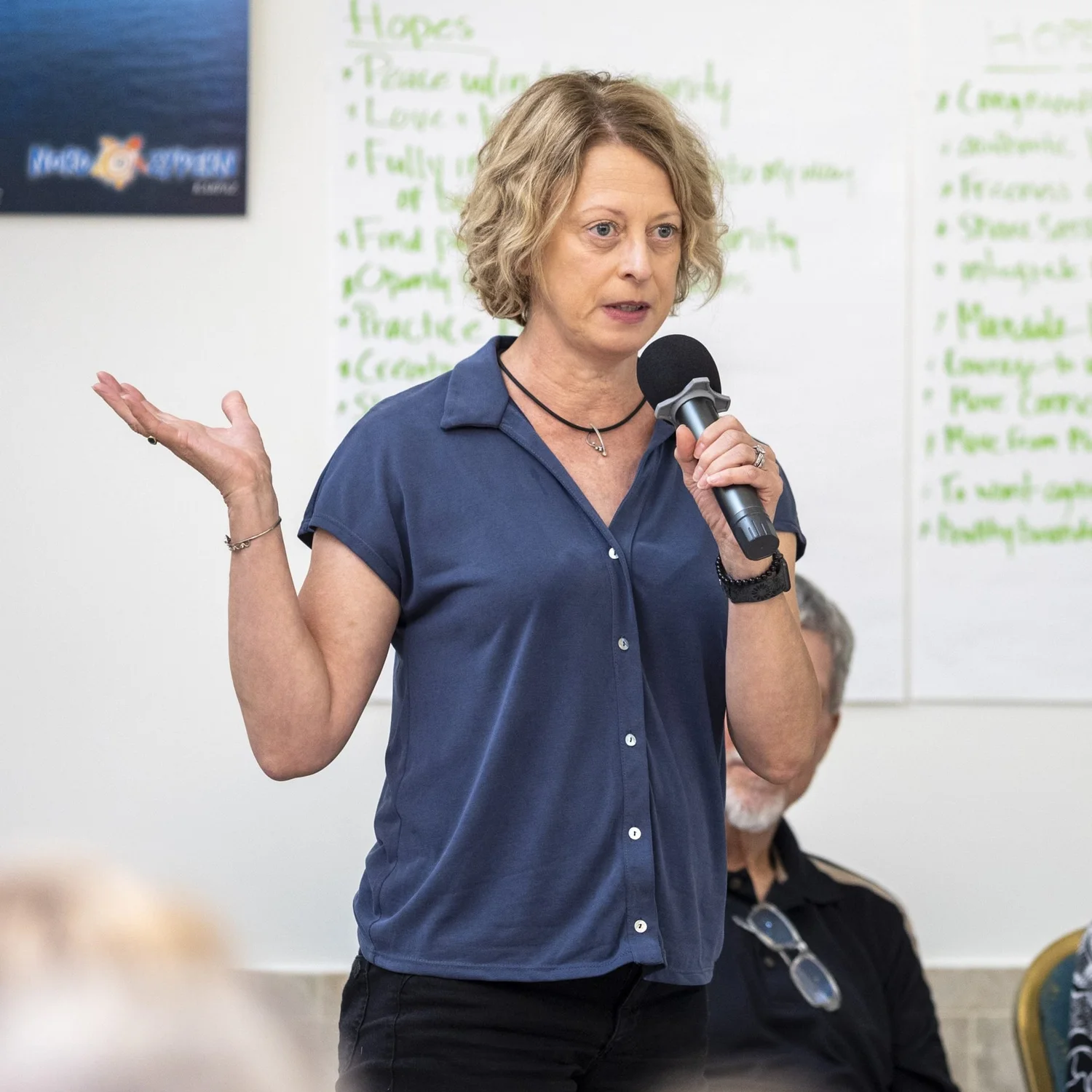 A woman in a navy shirt speaks into a microphone in front of a flip chart, one hand open as she talks
