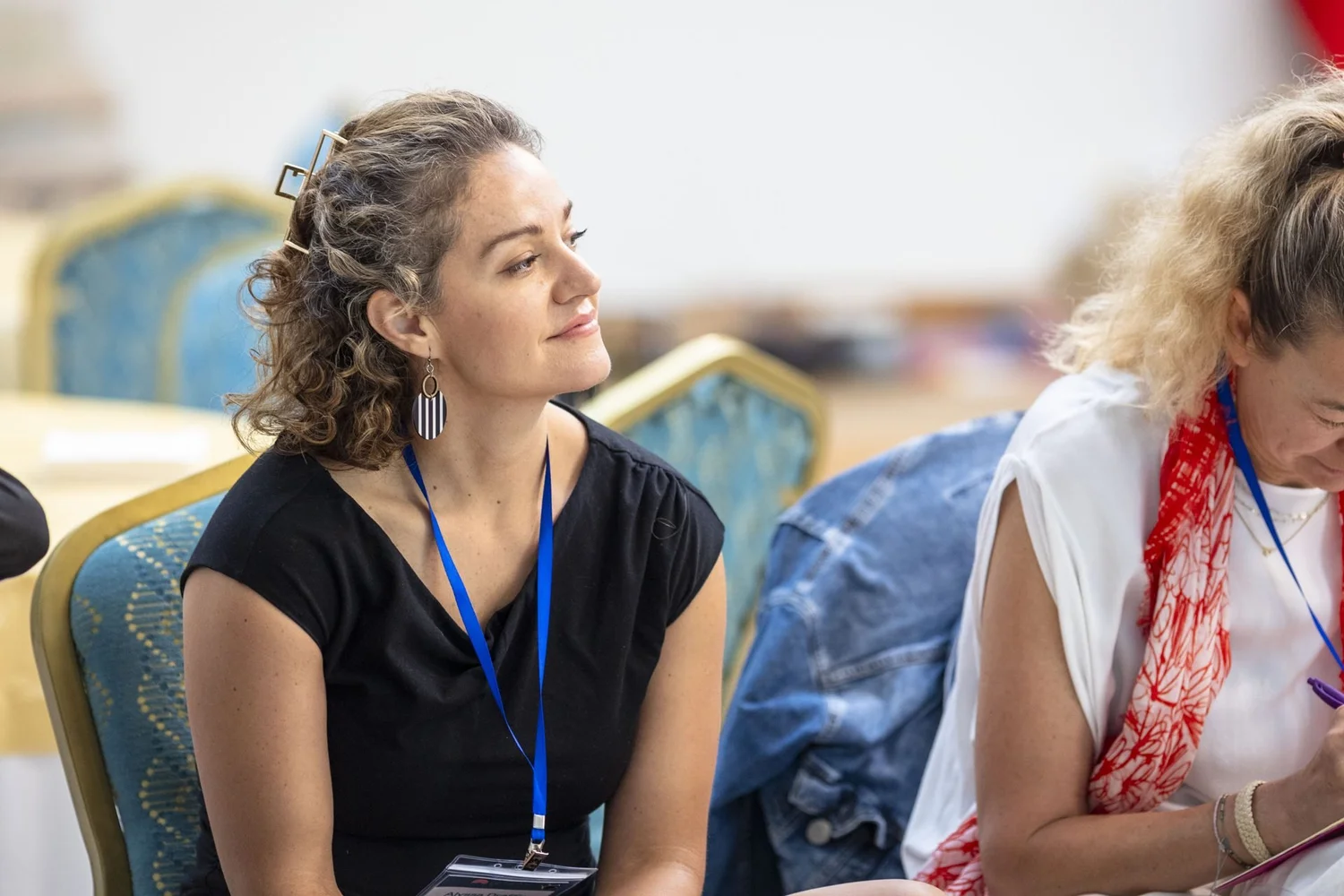 A woman with curly hair and a lanyard sits in a chair, looking ahead with a gentle smile