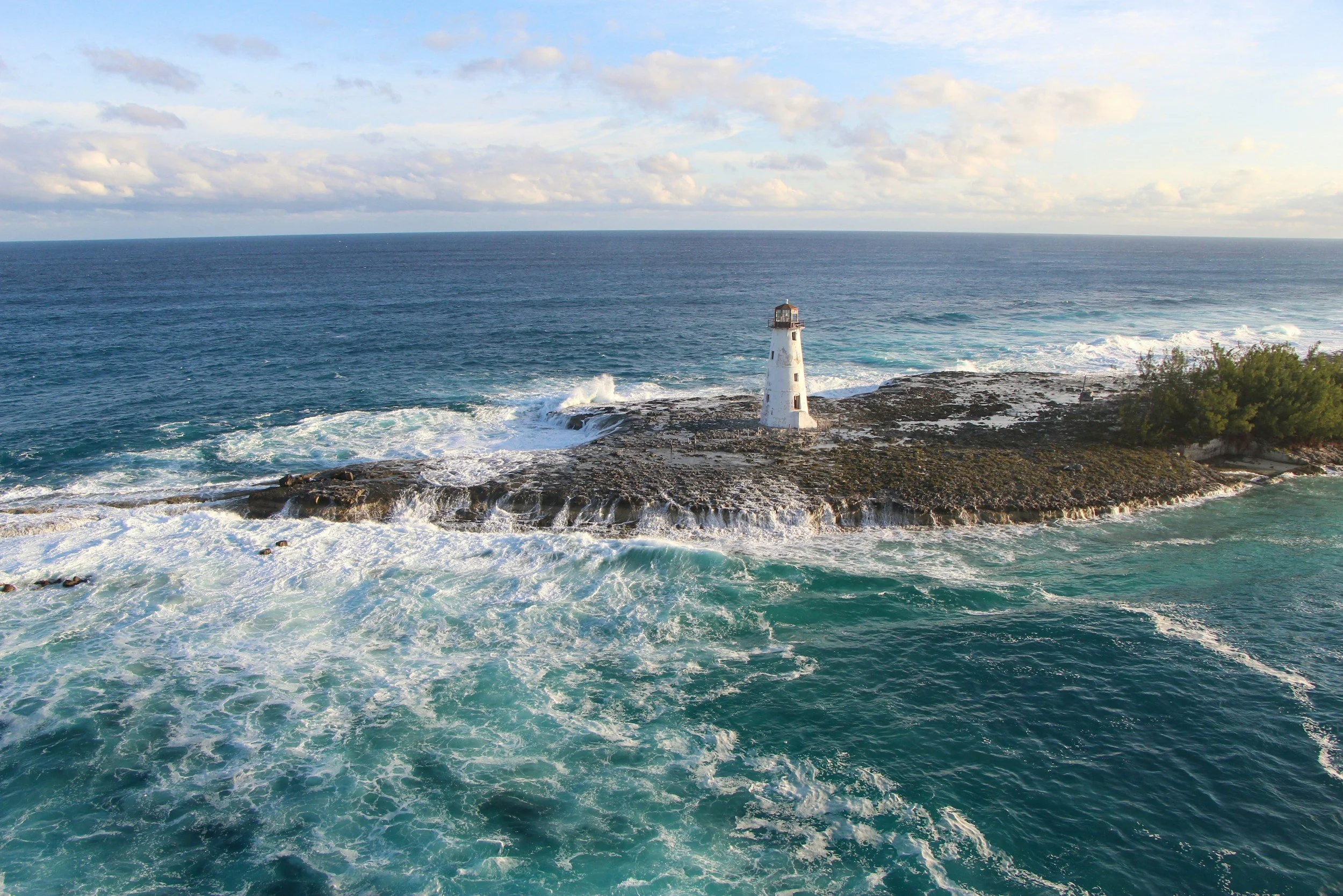 A lighthouse situated on rocky shoreline with waves crashing at its base, overlooking a vast ocean under a partly cloudy sky.