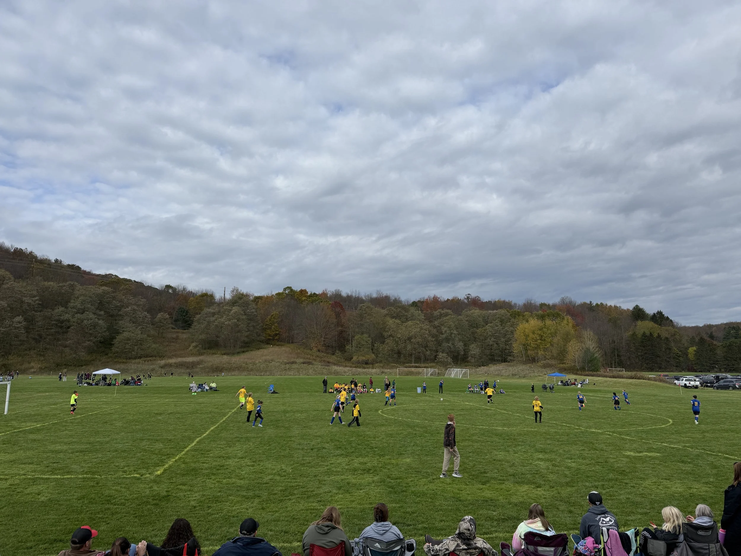 A soccer game taking place on a green field with players in yellow and blue jerseys, spectators watching from the sidelines, tents set up, and a forested hillside in the background under a cloudy sky.
