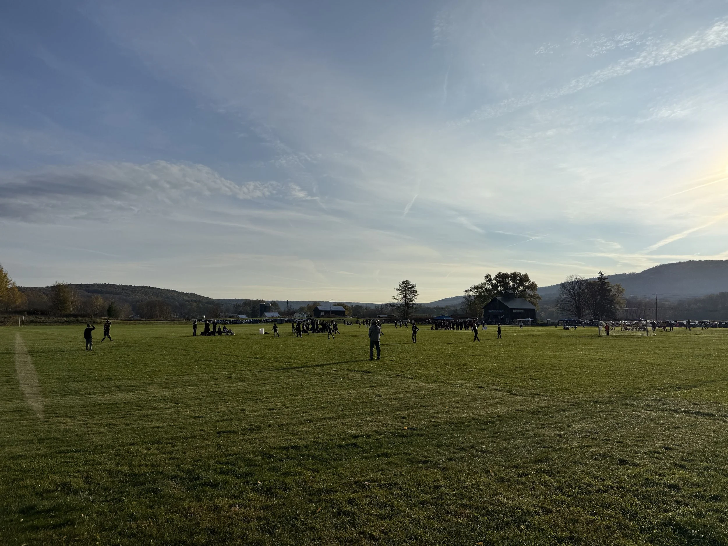 A wide open grassy field with people and horses, with distant hills and a partly cloudy sky at sunset.