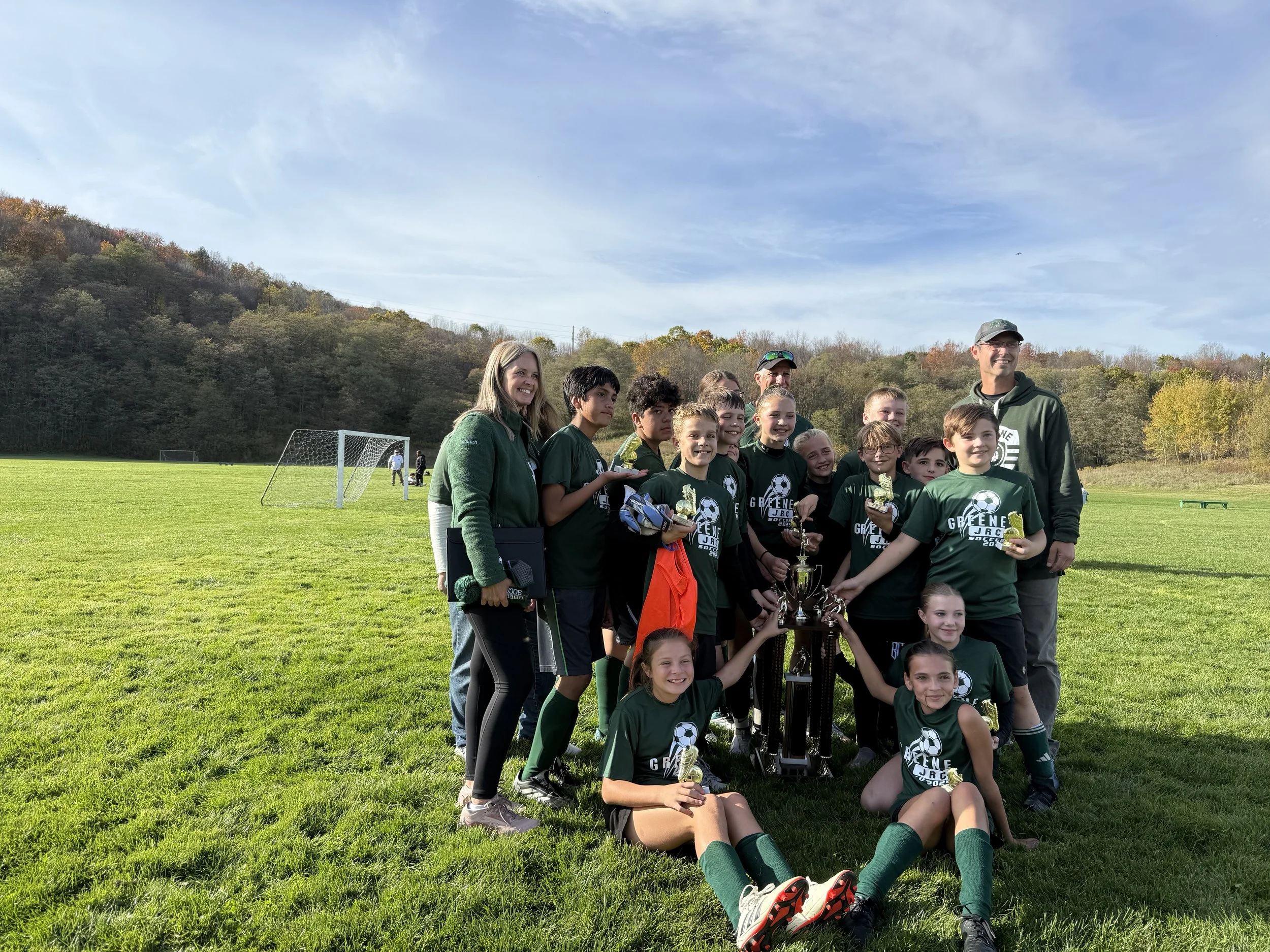 A youth soccer team in green jerseys and shorts, along with their coaches, celebrating on a grassy field with trophies. The sky is blue with some clouds, and there are trees in the background with fall foliage.