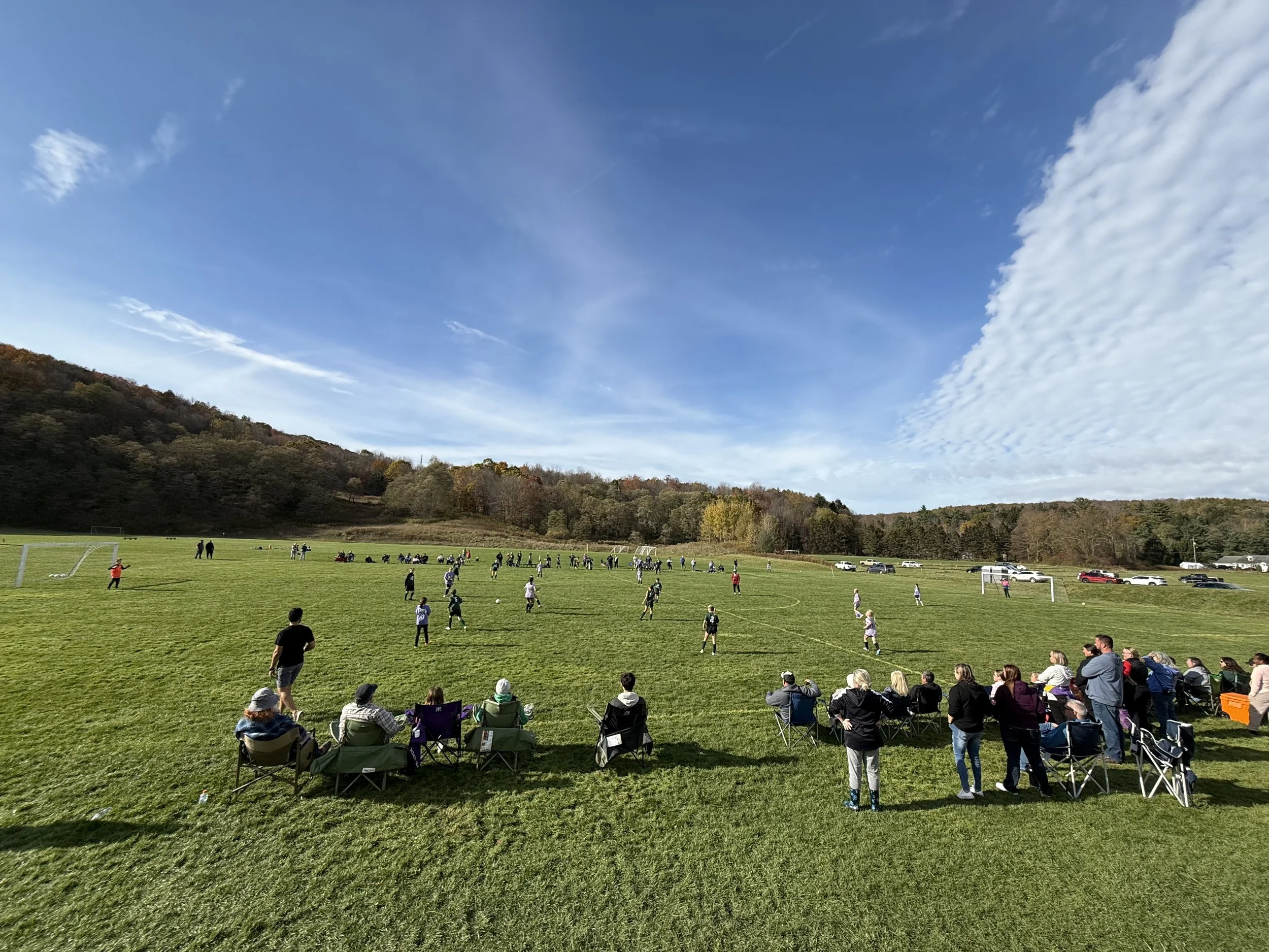 A group of people cheering and watching a soccer game on a grassy field under a partly cloudy sky, with some spectators sitting in foldable chairs and others standing along the sidelines, surrounded by cars parked in the distance.