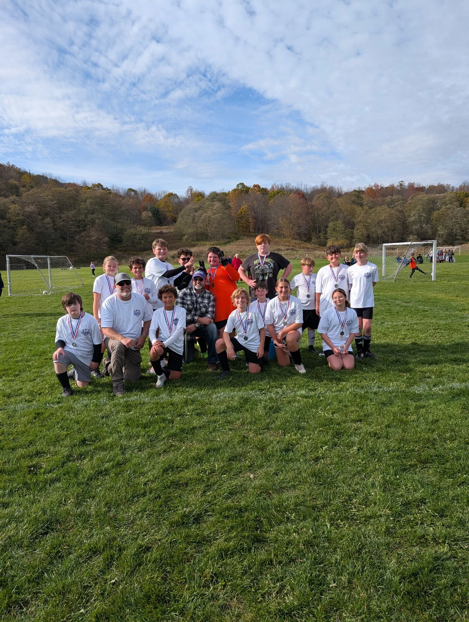 Group of children's soccer team with medals posing on a soccer field with trophies, coaches, and teammates in the background, under a partly cloudy sky.