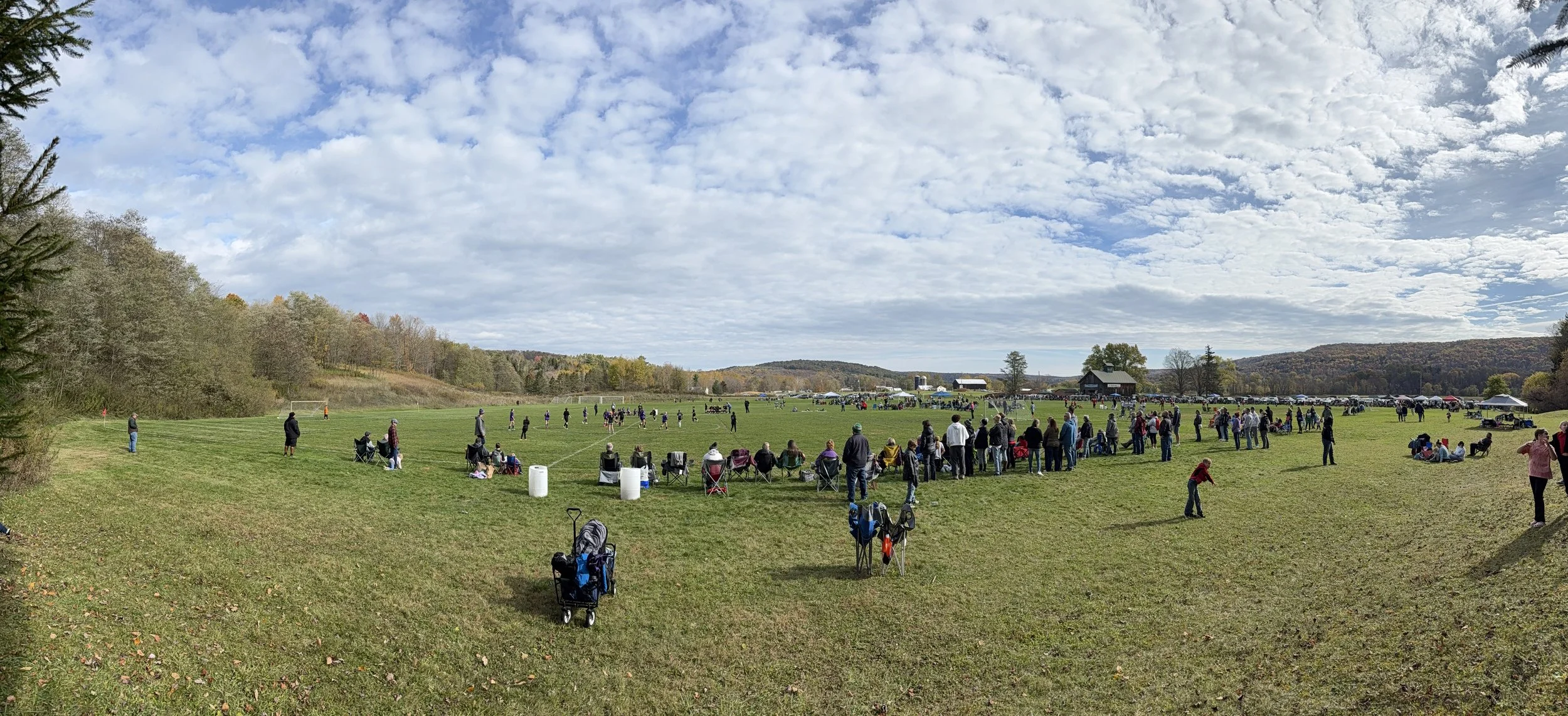 Outdoor soccer game on a large grassy field with spectators watching and sitting in chairs, surrounded by trees and hills under a partly cloudy sky.