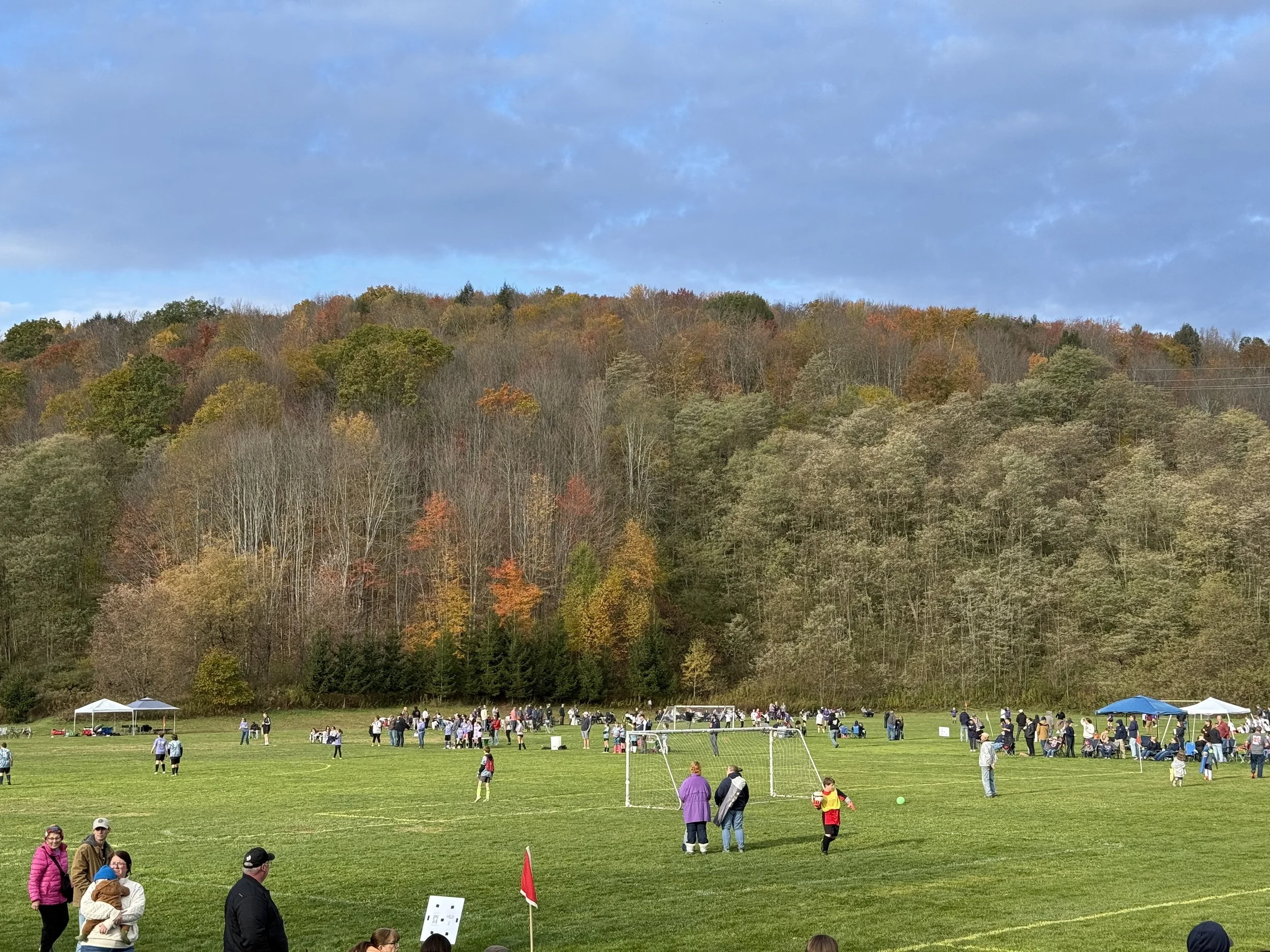 A large open grassy field with people gathered for outdoor activities, some near small soccer goals and tents, with a wooded hillside in autumn colors in the background under a partly cloudy sky.