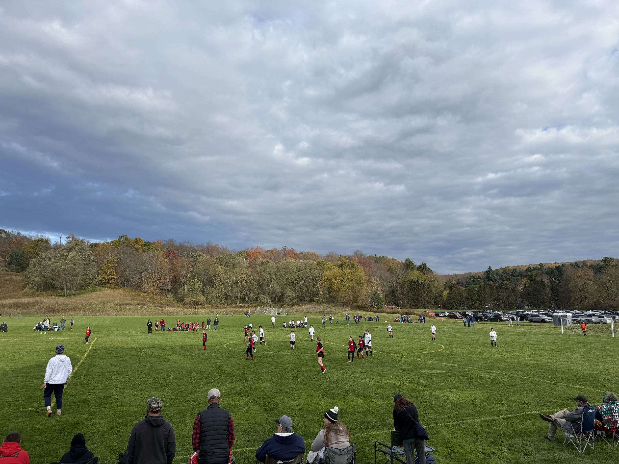 A soccer game is taking place on a large grassy field with players in red and white uniforms. Spectators are watching from the sidelines, and there are trees with fall foliage in the background under a cloudy sky. There are also parked cars on the ri