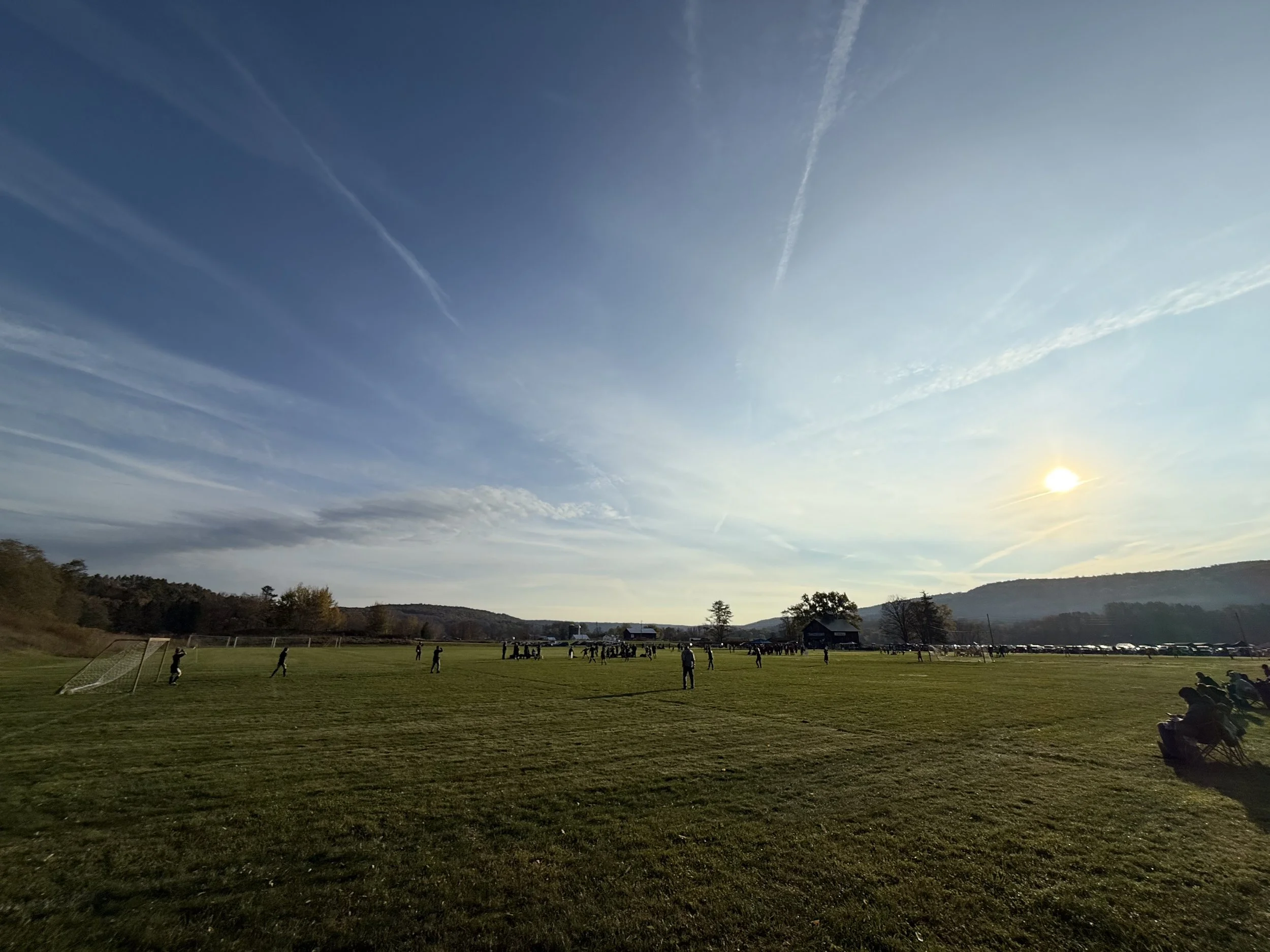 A grassy field with people playing soccer, surrounded by mountains and trees, under a partly cloudy sky with the sun shining.