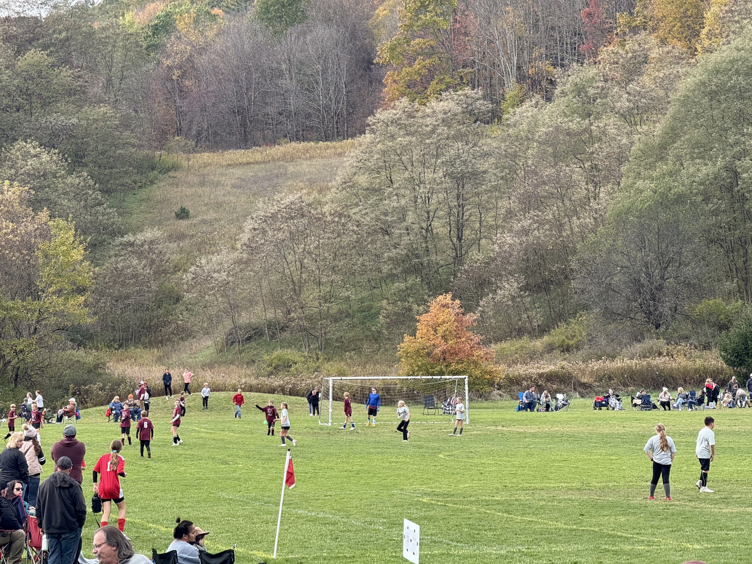 A soccer game is occurring on a grassy field, with players in maroon and white uniforms. Spectators are gathered around, watching and sitting on chairs. Trees and a hillside with autumn foliage are in the background.