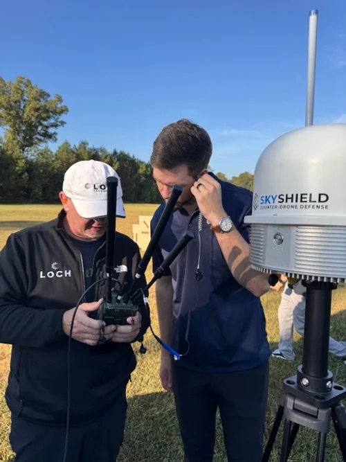 Two men operating a SkyShield drone defense system outdoors near Ft. Benning testing drone detection and jamming capabilities for the ARMY on a grassy field, with trees and a clear blue sky in the background.