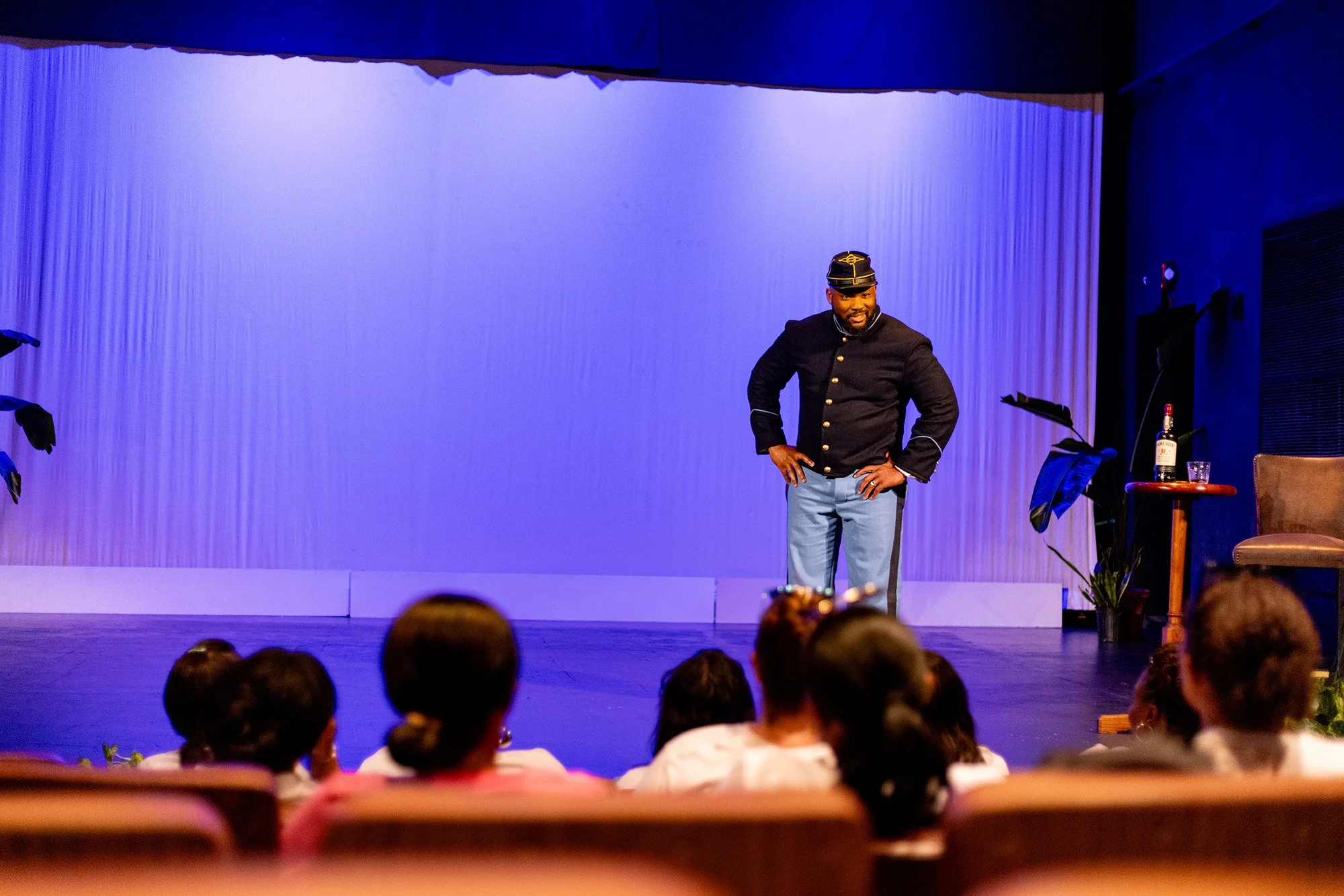 A man in a vintage black uniform and hat on stage with an audience in front, with a blue-lit backdrop and a side table with a bottle and glass.