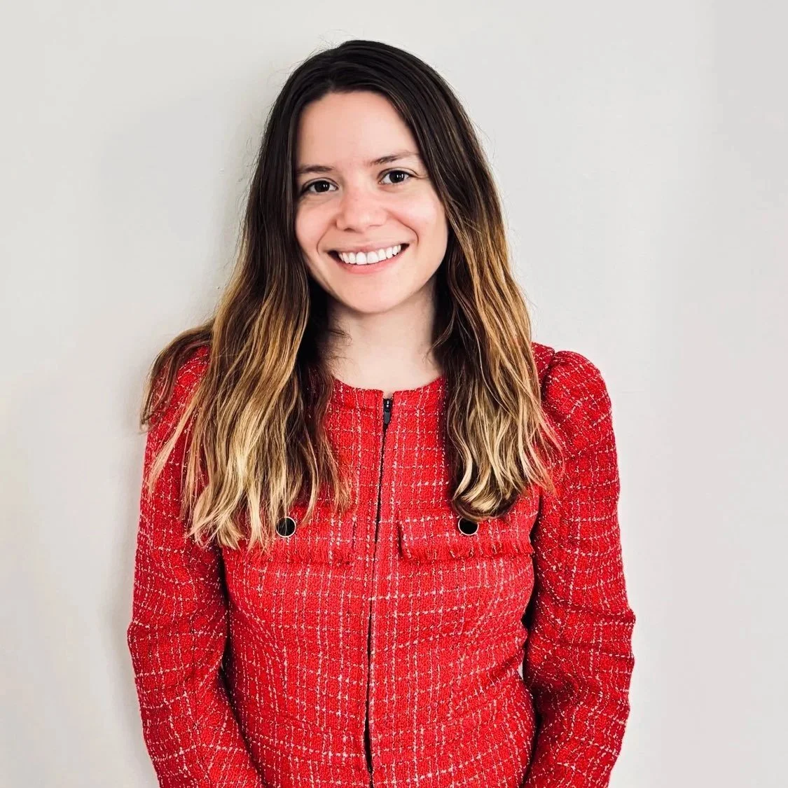 Young woman with long brown hair smiling, wearing a red textured jacket, standing against a plain white wall.