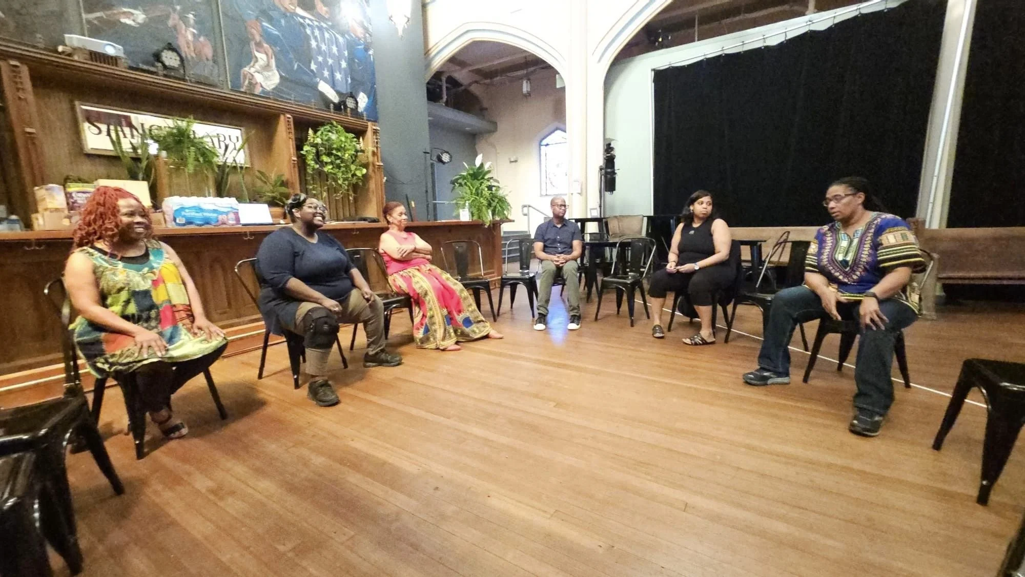 Six people are sitting in a semi-circle inside a room with wooden floors and stage. Five women and one man, all appear to be part of a discussion or workshop.