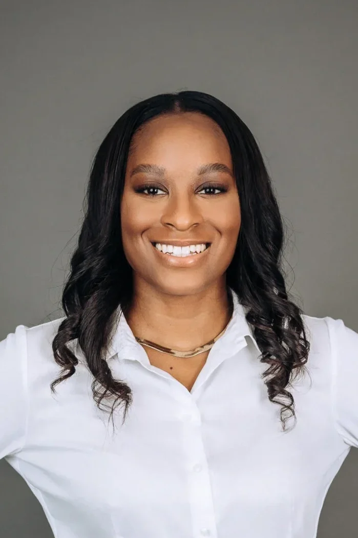 A young Asian woman with shoulder-length black hair smiling outdoors with green foliage in the background.