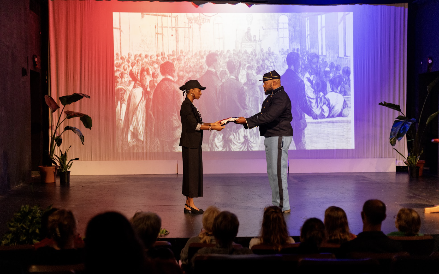Two people on stage exchanging a certificate or award, with a black and white historical photo projected on the background, and an audience watching.