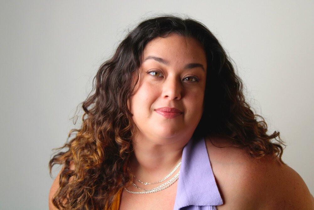 A woman with dark hair pulled back, wearing a white shirt and black vest, standing in front of red curtains.