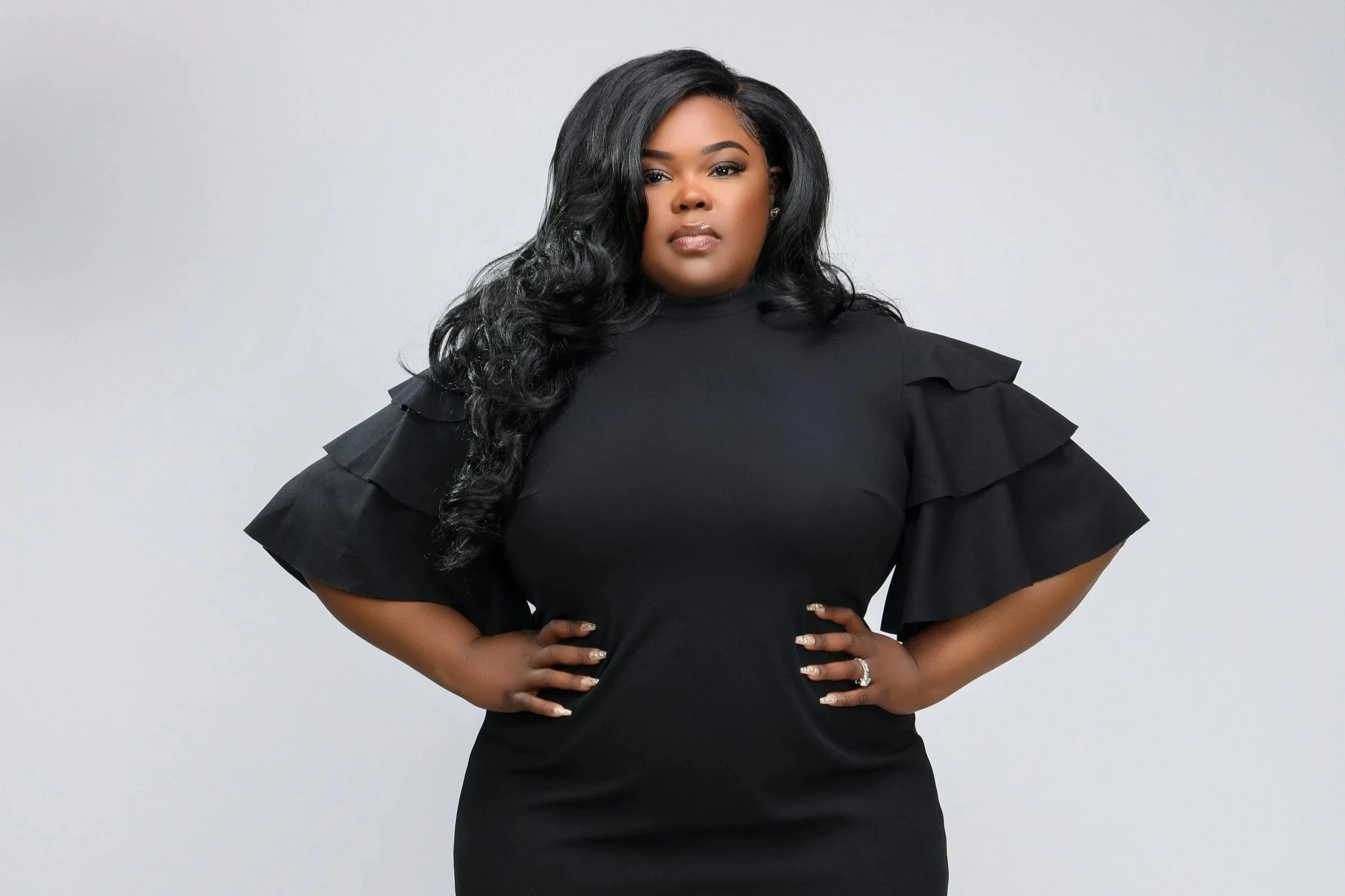 A confident woman with her hands on her hips, wearing a black dress with ruffled sleeves and clear heels, standing against a plain background.