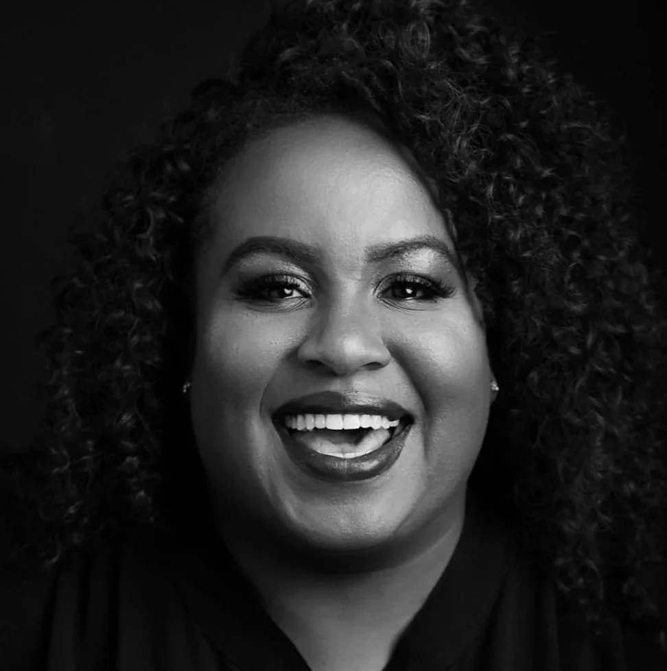 Woman in a black blazer and tulle skirt smiling, standing against a white background with hands on hips.
