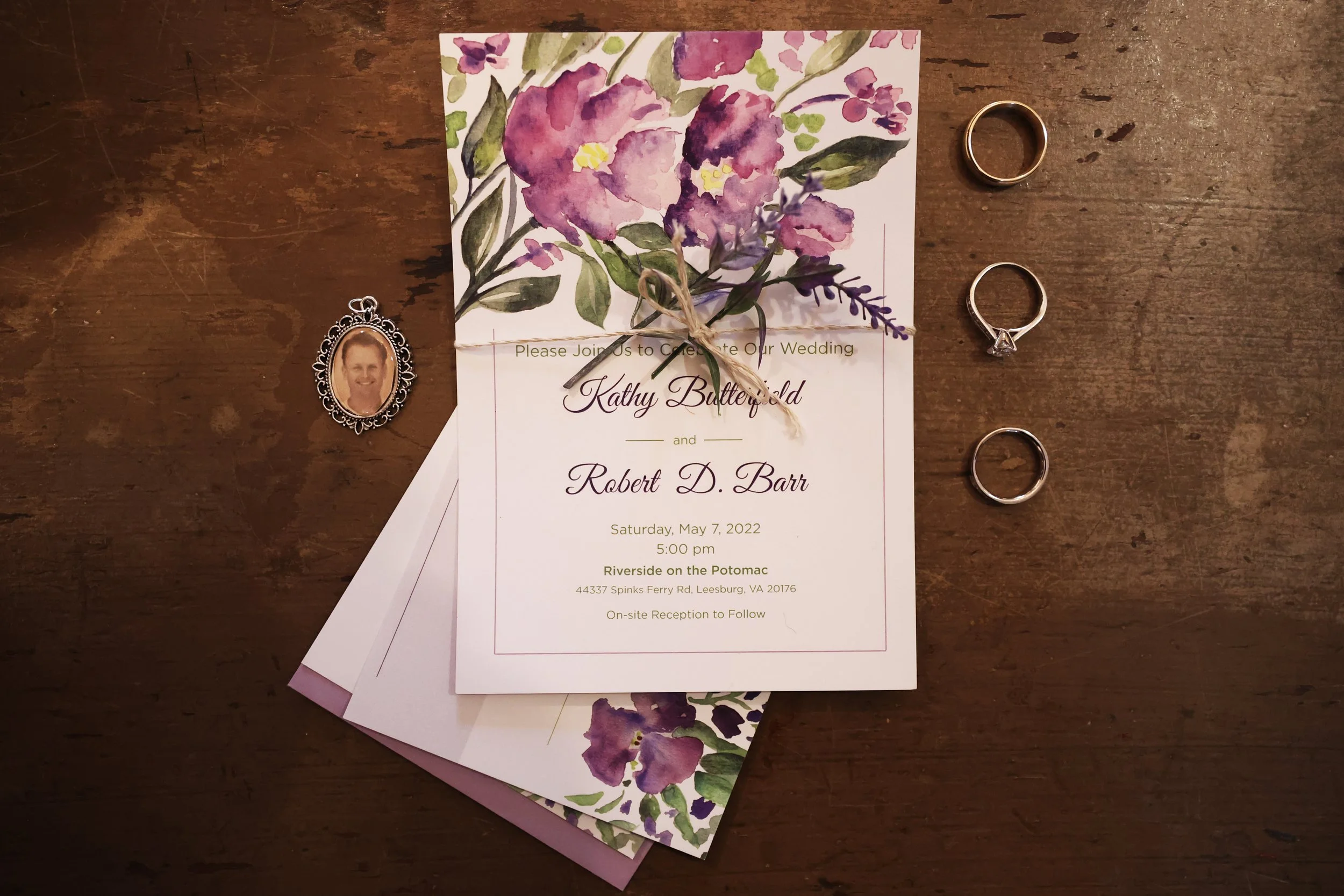 A wedding invitation with purple floral design, three rings, and a photo pendant on a wooden surface.