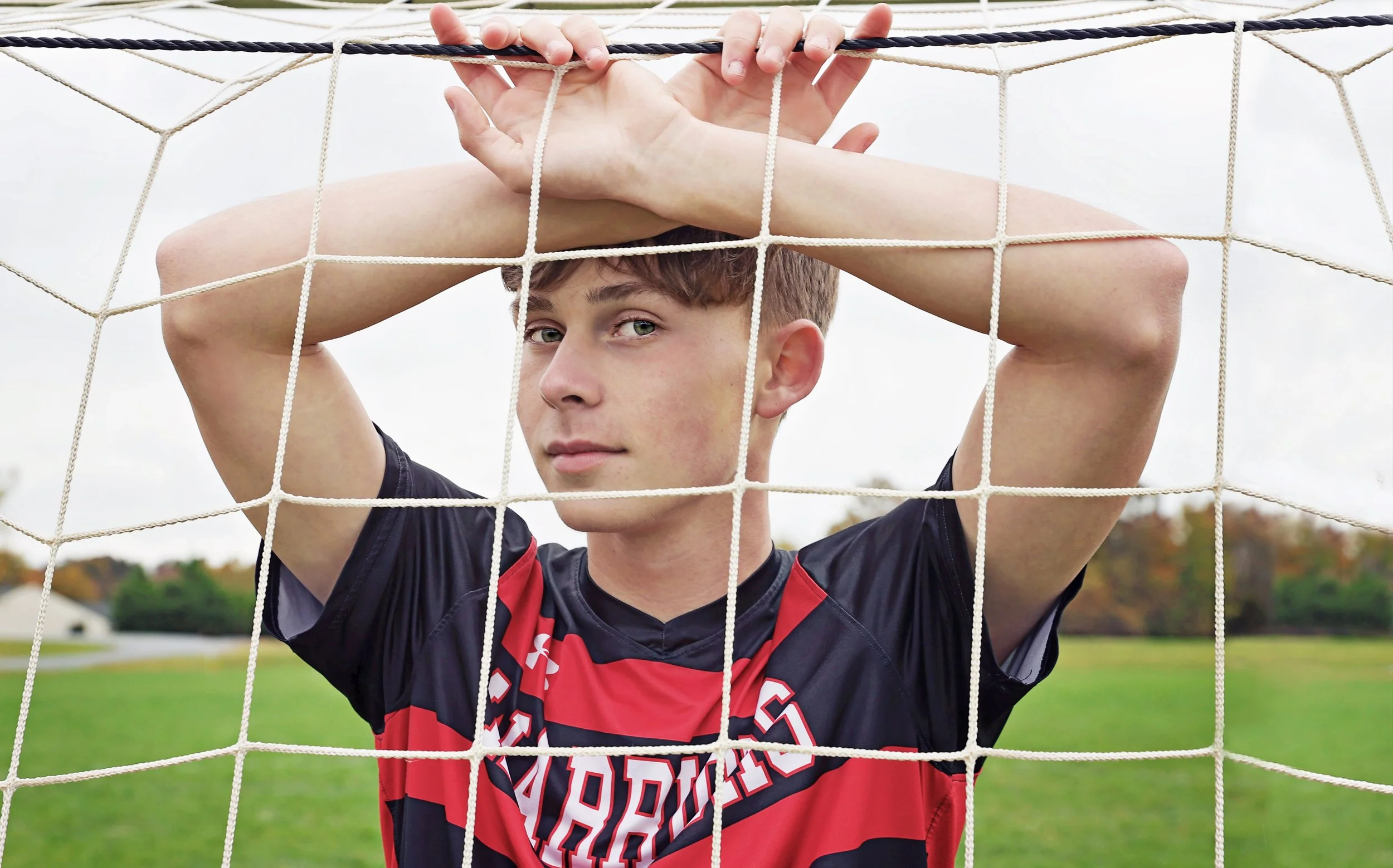 Young male soccer player in a black and red uniform leaning against a soccer goal net, outdoors on a grassy field with trees in the background.