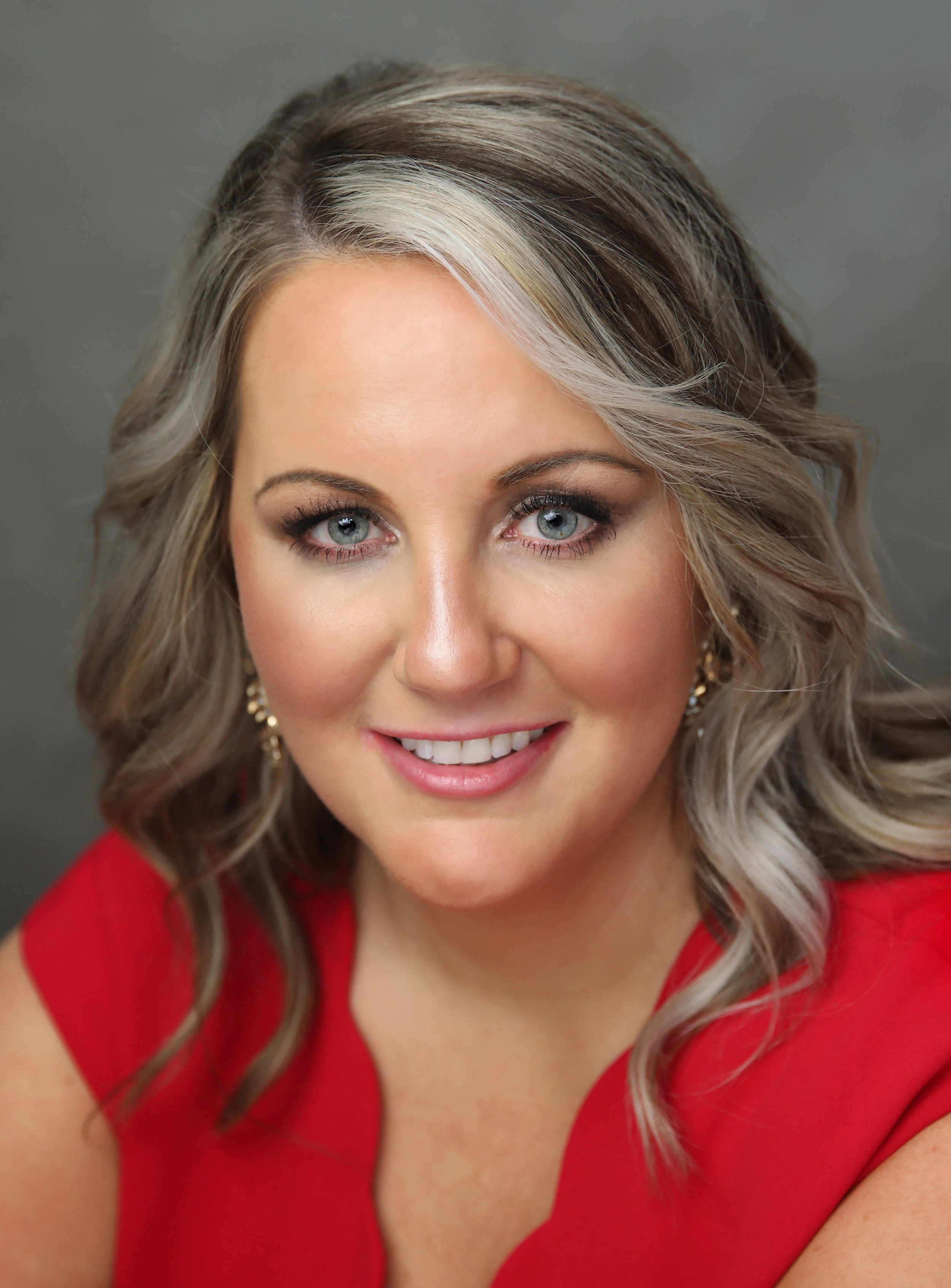 A woman with blonde, wavy hair and blue eyes, wearing makeup, earrings, and a red top, smiling at the camera against a gray background.