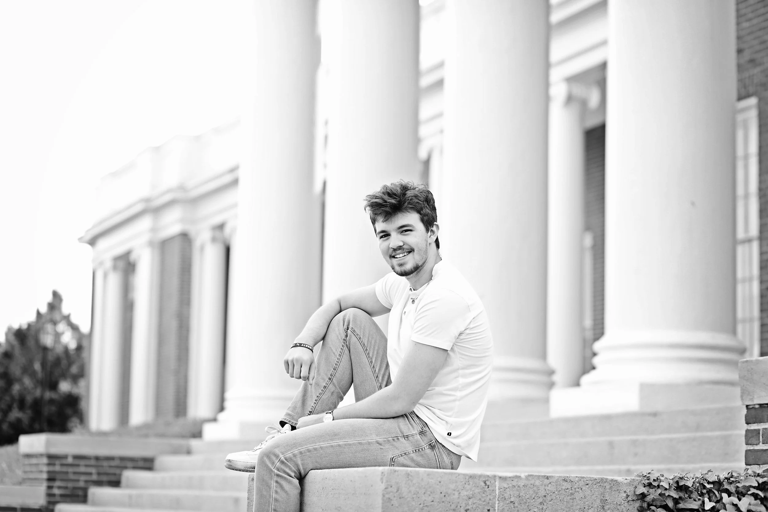 A young man sitting on steps outside a building with large columns, smiling towards the camera, in black and white.
