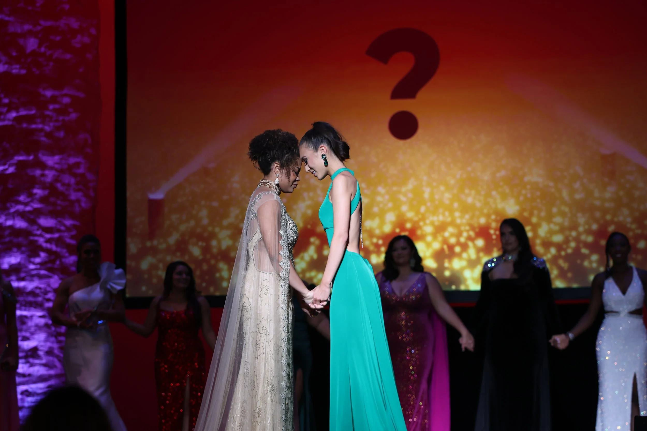 Two women holding hands and touching foreheads on stage during a pageant or contest, with other women in evening gowns in the background.