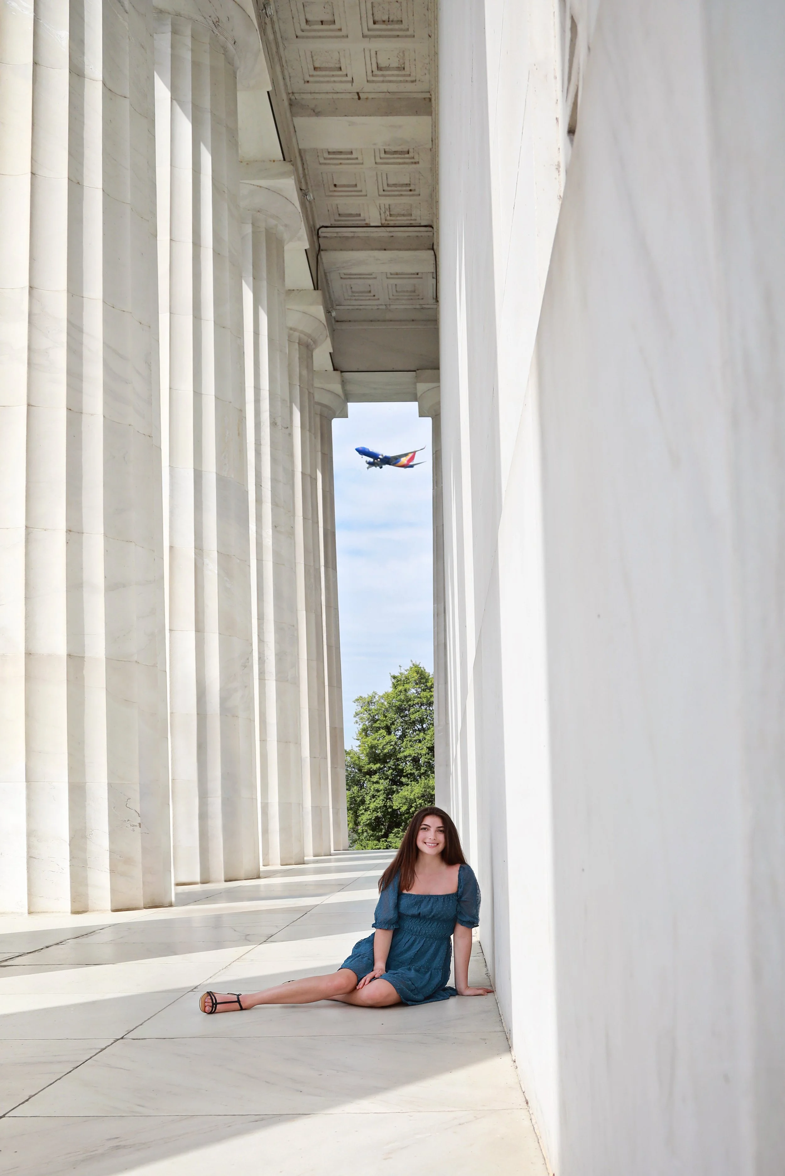 A young woman in a blue dress sits on the ground in front of a white marble monument with tall columns, with an airplane flying overhead in the sky.