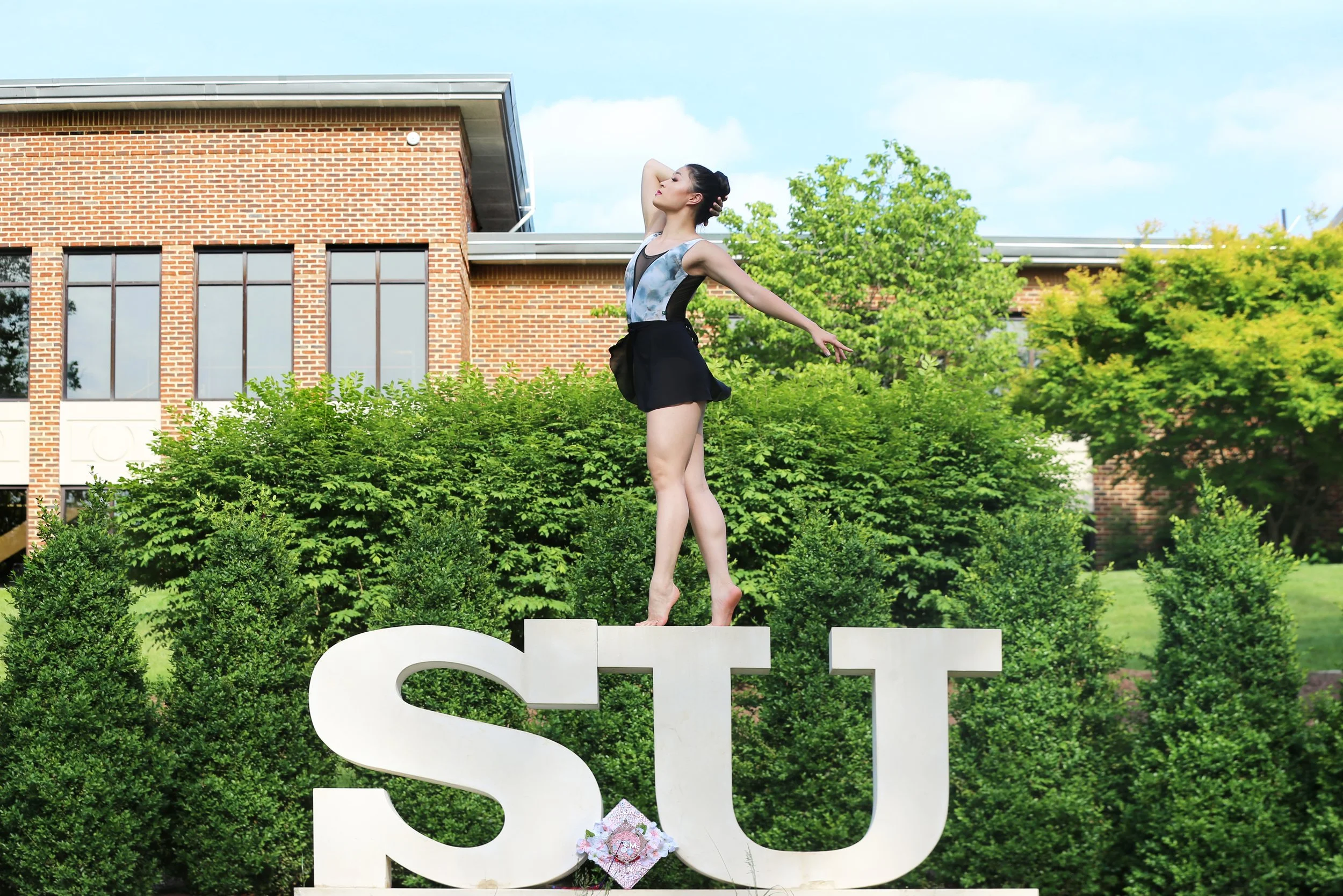A young woman in a dance or gymnastics outfit standing on large white letters spelling 'SU' outdoors, with greenery and a brick building in the background.