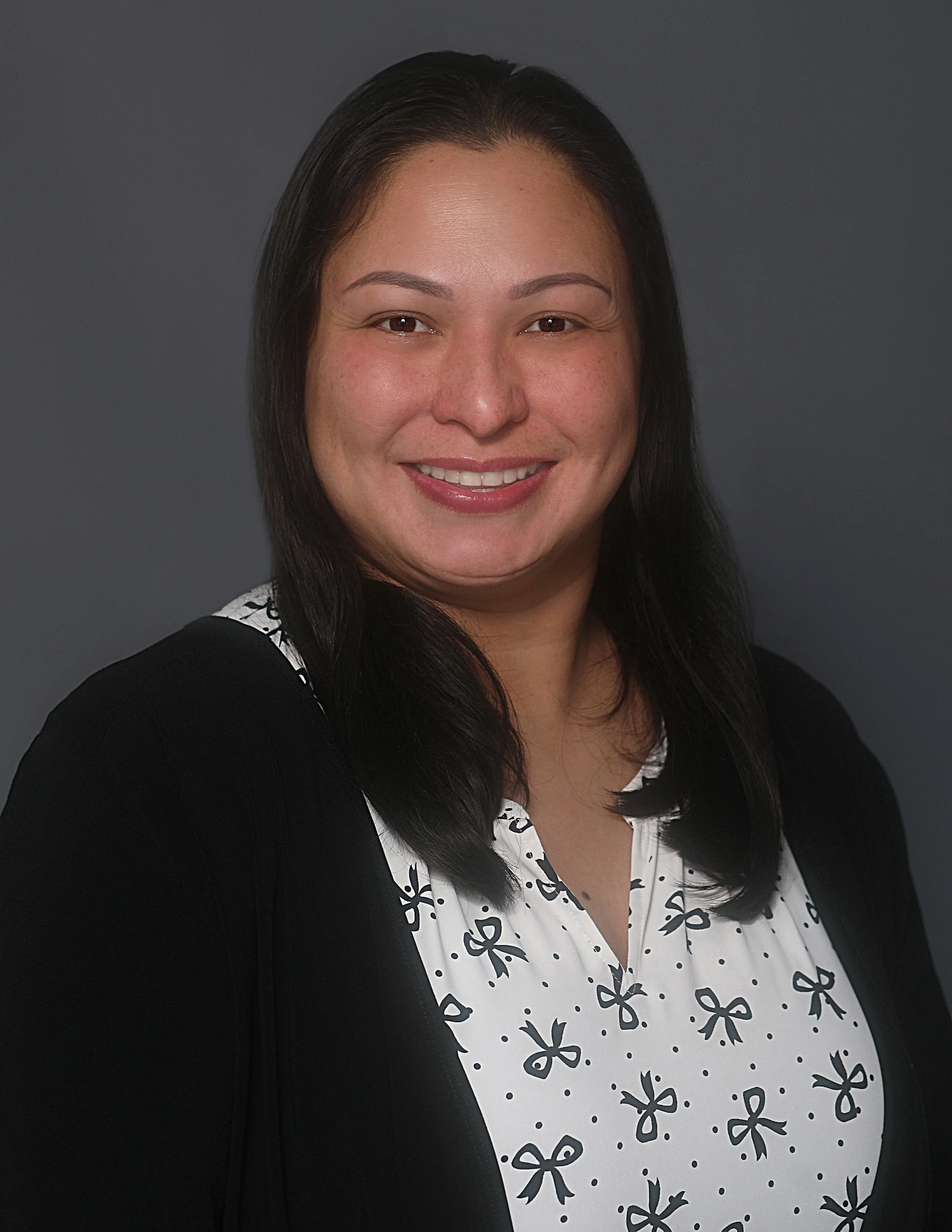 Professional headshot of a woman with shoulder-length black hair, smiling, wearing a black blazer over a white blouse with black bow patterns, against a dark gray background.