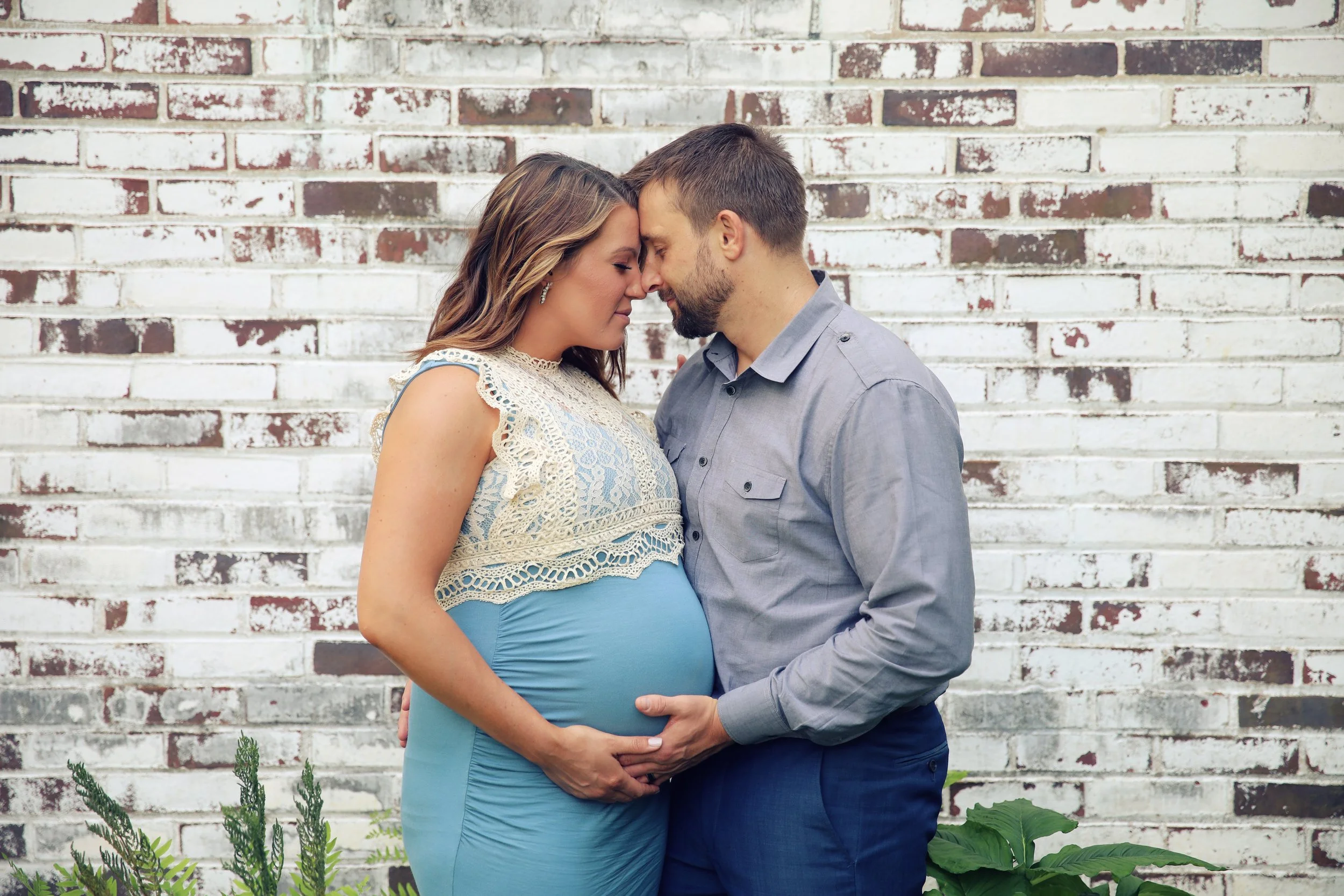 A pregnant woman and her partner standing close together with foreheads touching against a white brick wall, with greenery in the foreground.