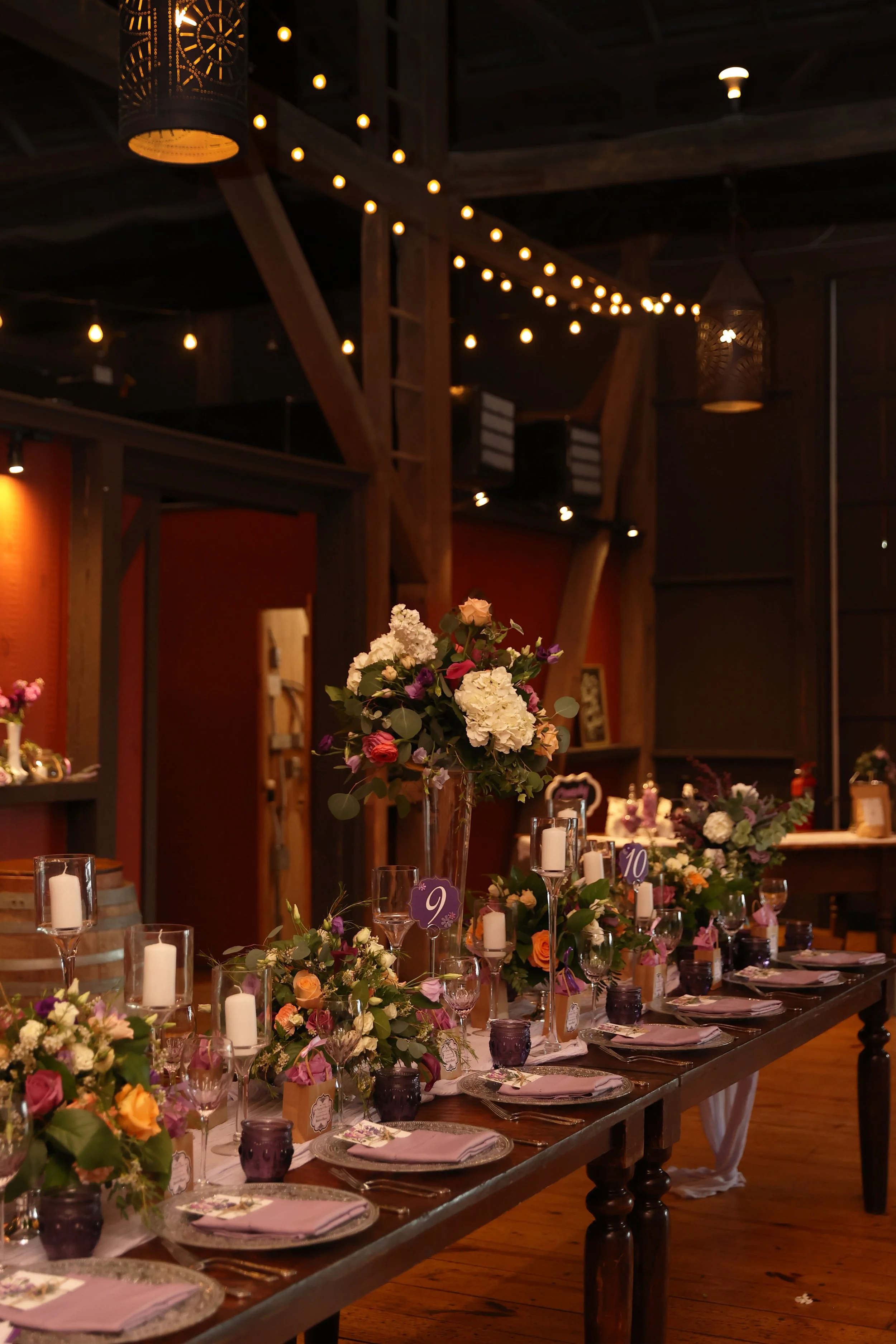 Wedding reception table decorated with floral centerpieces, candles, and tableware in a rustic venue with string lights.