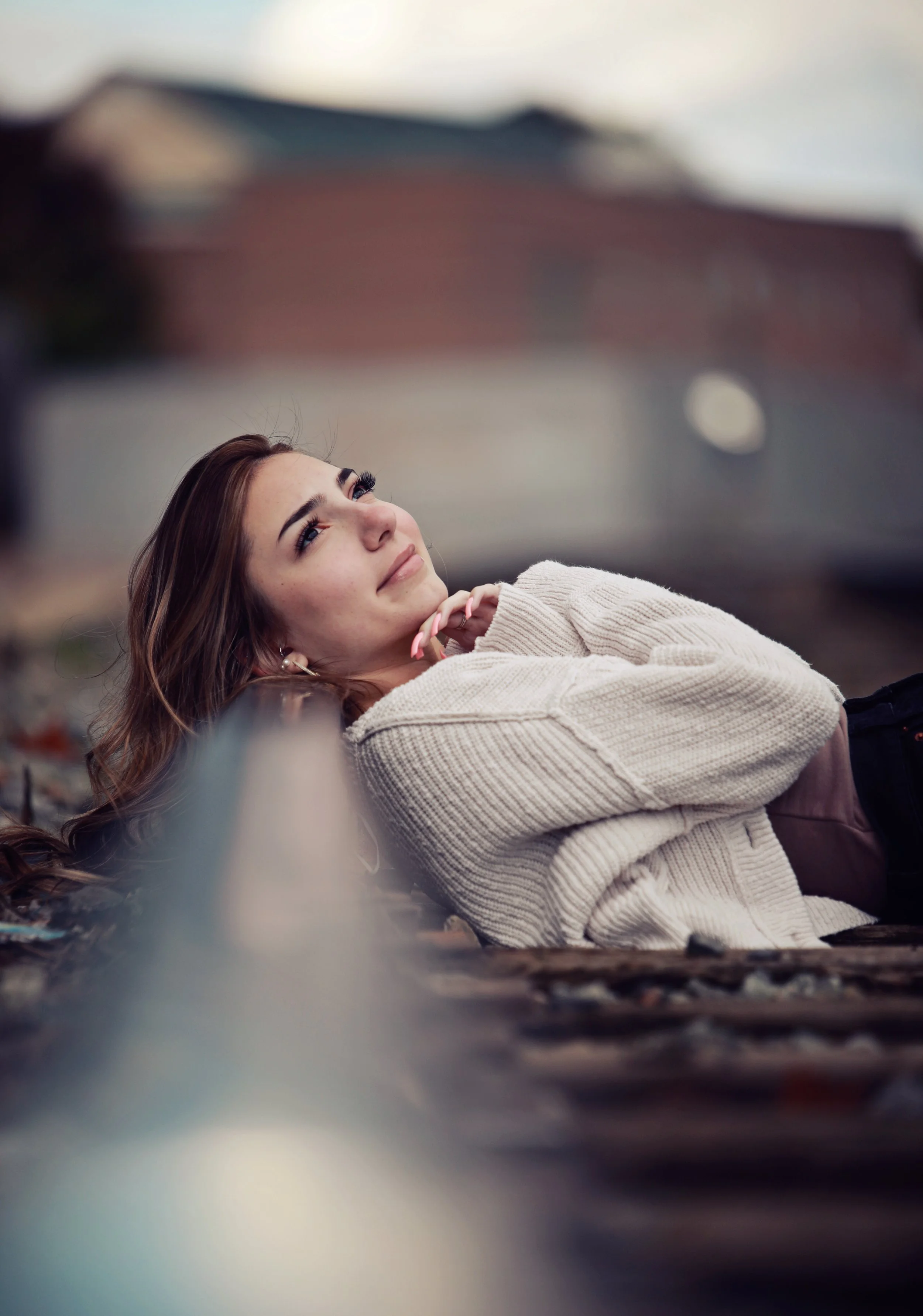 A young woman with brown hair and dark eyebrows laying down on her side on railway tracks, wearing a beige sweater and earrings, looking up with a calm expression.