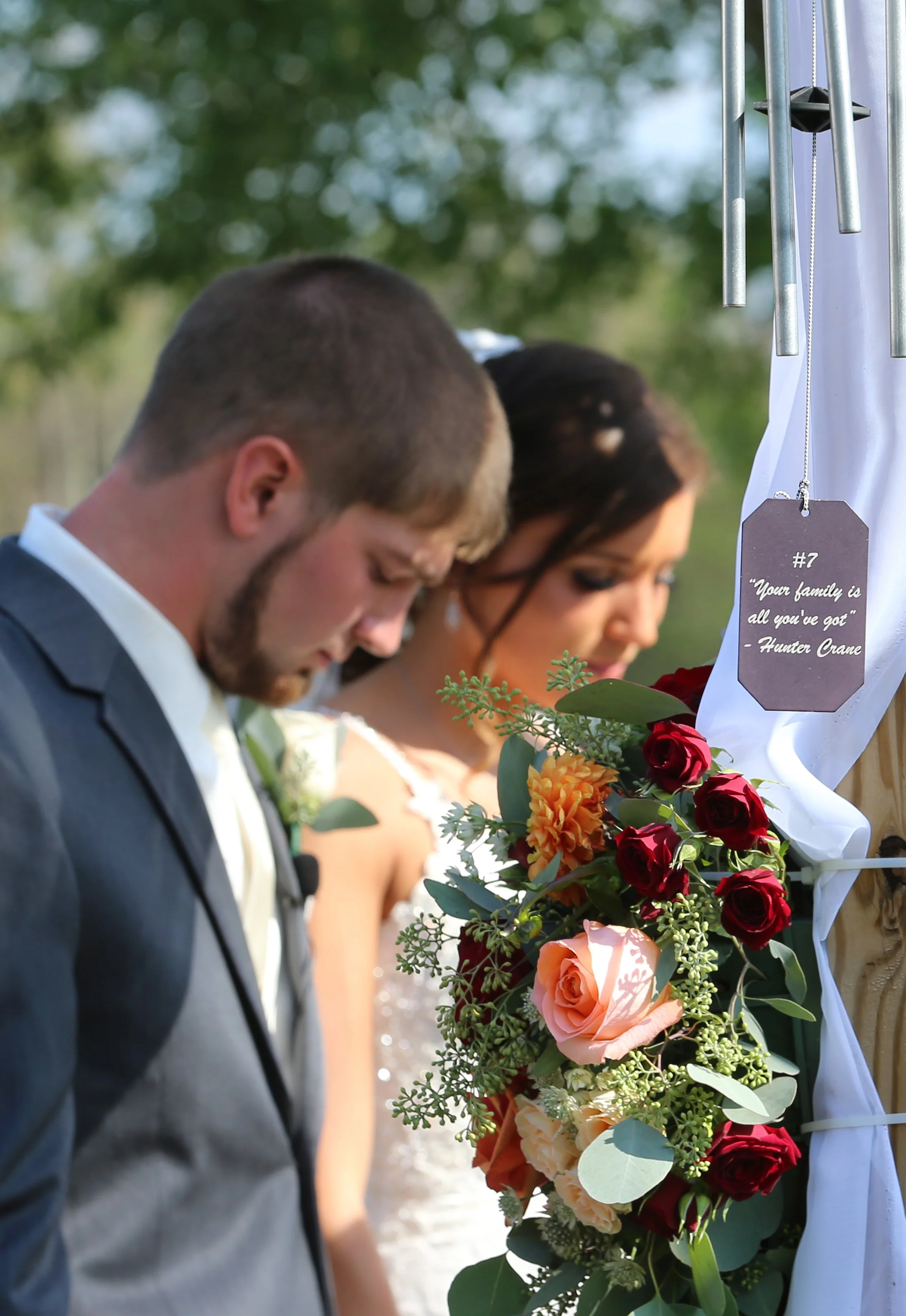 A bride and groom praying or holding their heads bowed during a wedding ceremony outdoors, with a floral arrangement of red and peach roses and greenery in front of them, a white ribbon and a sign that reads 'Your family is all you've got' by Hunter Crane.