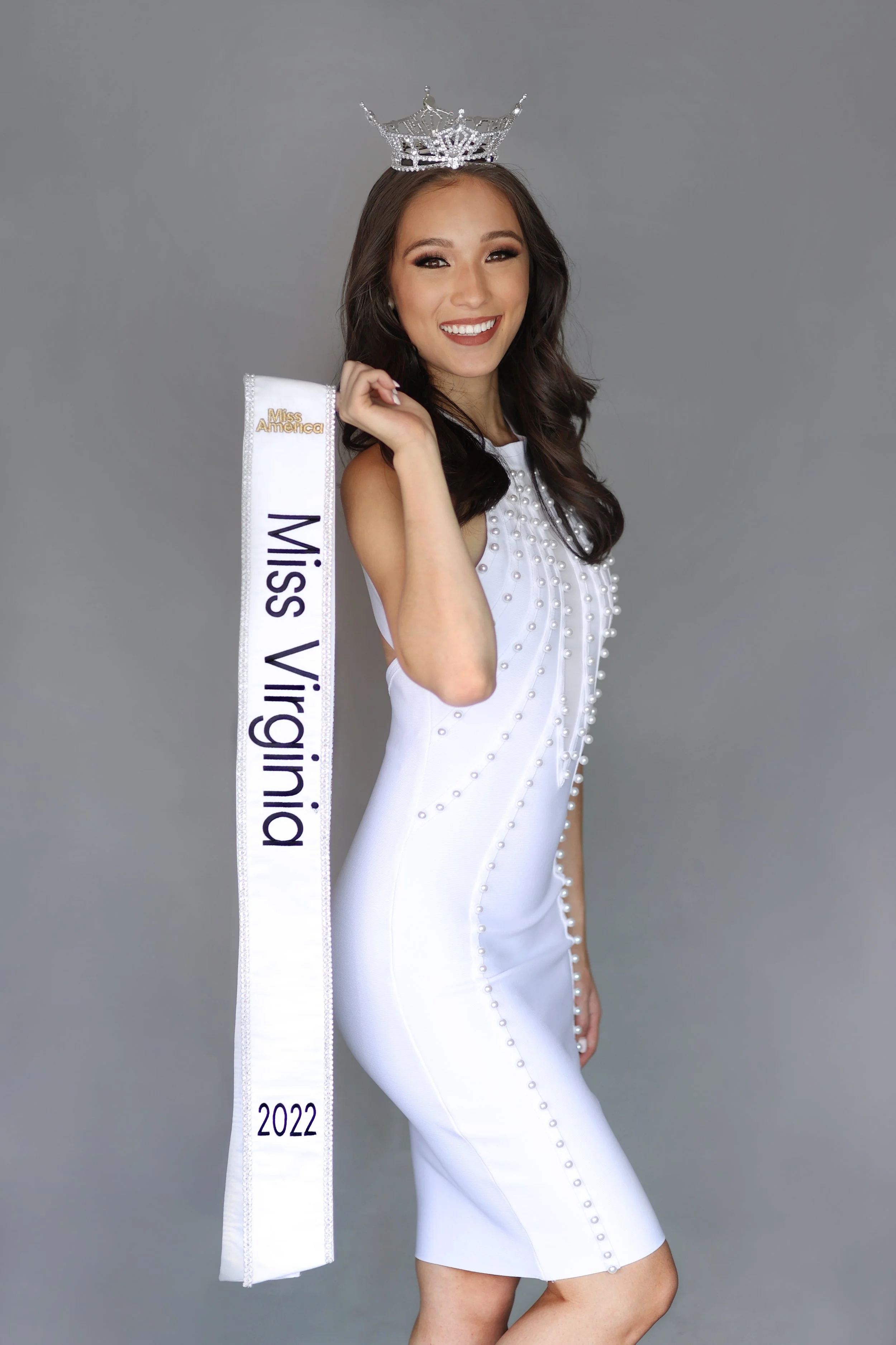 A woman wearing a white dress with pearl embellishments, a sash reading 'Miss Virginia' 2022, and a crown on her head, smiling and posing against a gray background.