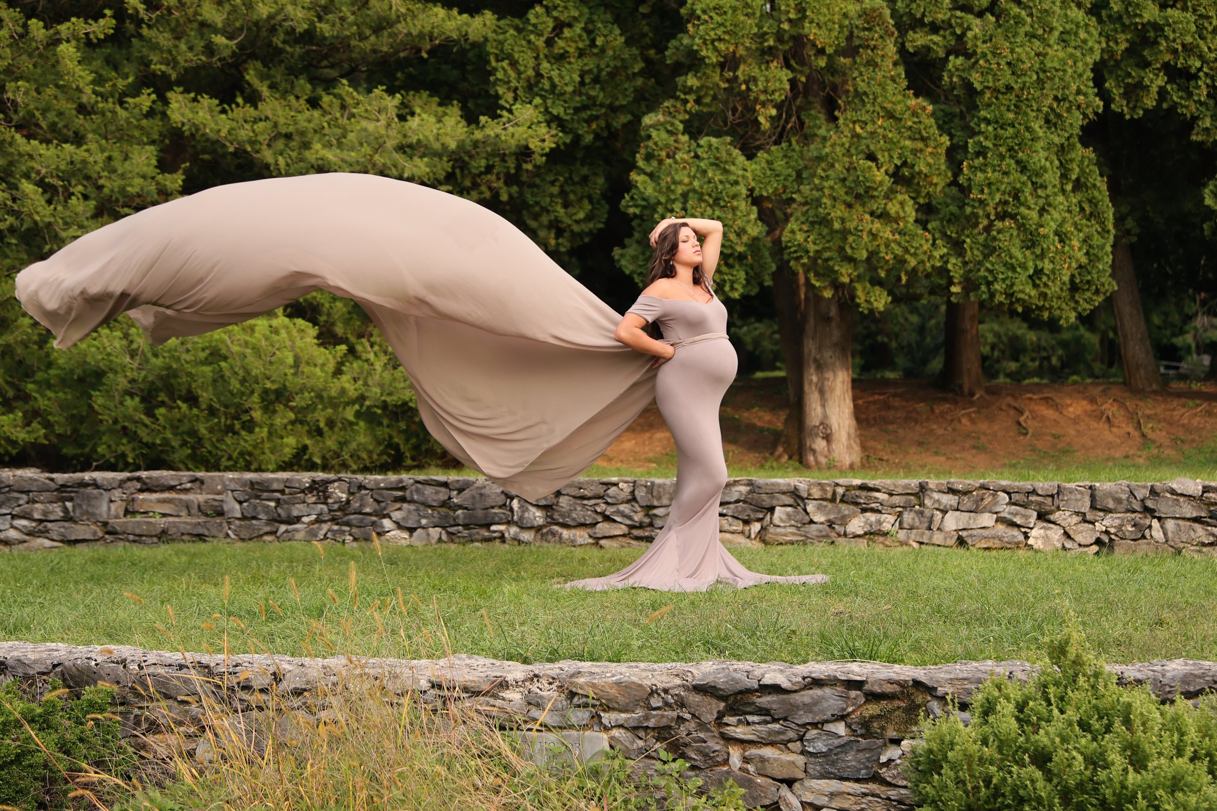 Pregnant woman in a flowing beige dress standing outdoors on grass with a stone wall and trees in the background.