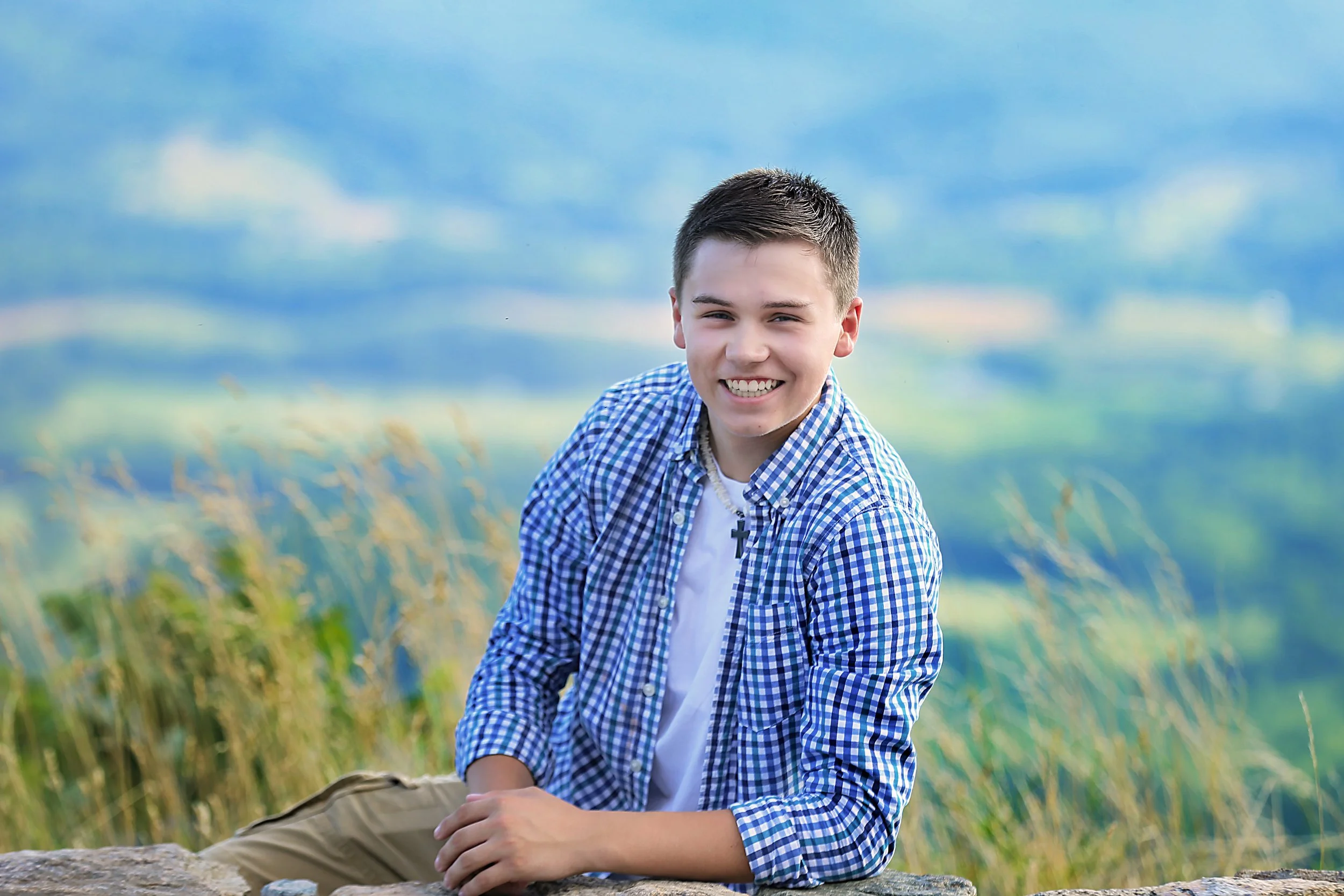 A young boy with short dark hair and a big smile, wearing a blue checkered shirt and tan pants, leaning on a rock outdoors with a blurred scenic background of blue sky and green hills.