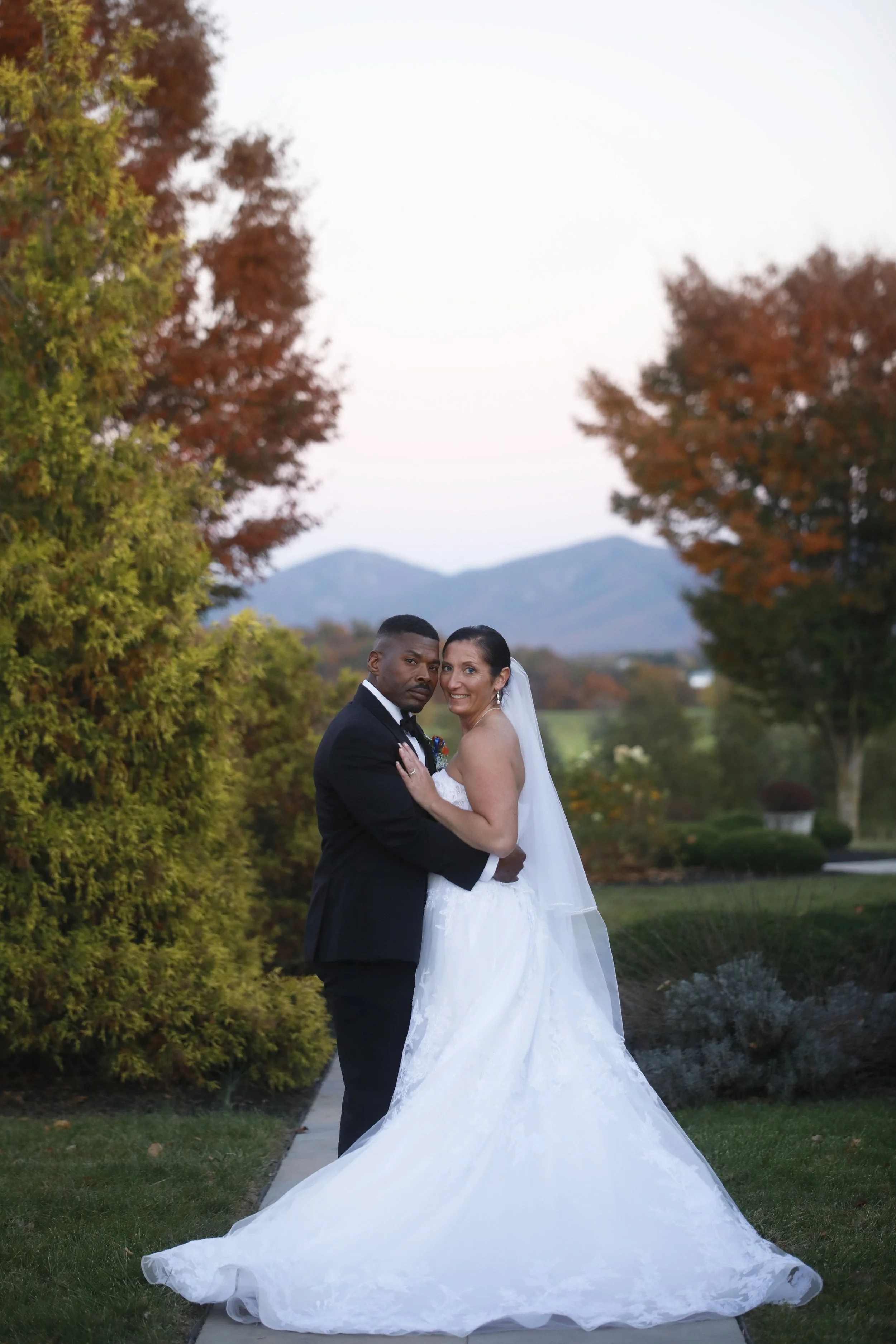 A bride and groom standing outdoors in a park-like setting with autumn trees and mountains in the background during sunset.