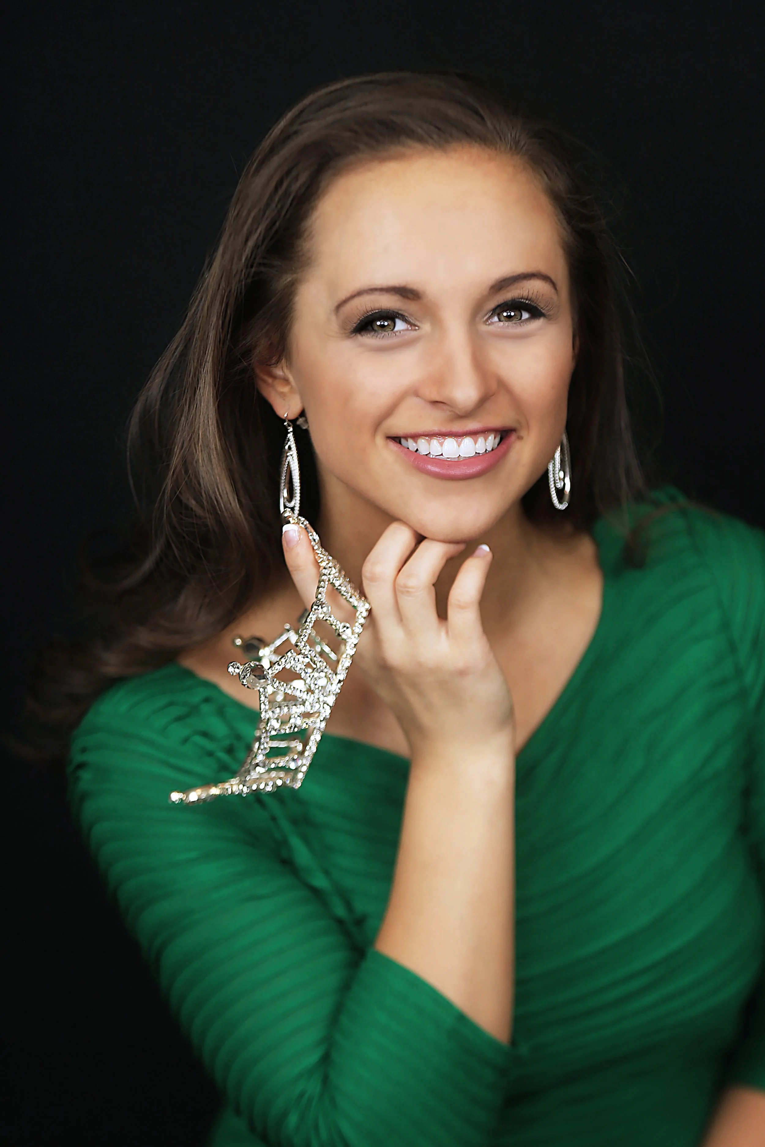 Portrait of a smiling woman with brown hair, wearing a green dress and silver jewelry, posed against a black background.