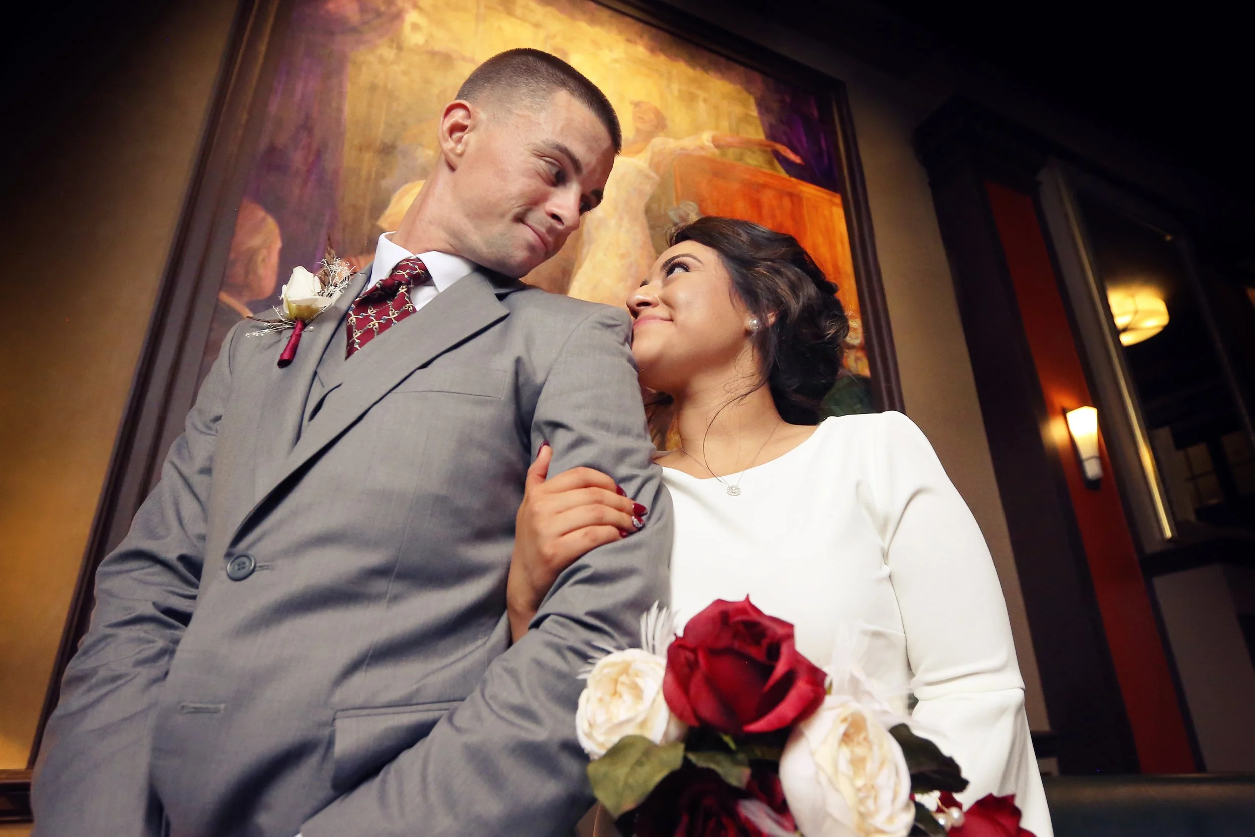 A bride and groom looking at each other affectionately inside a hotel or restaurant, with the bride holding a bouquet of red and white roses.