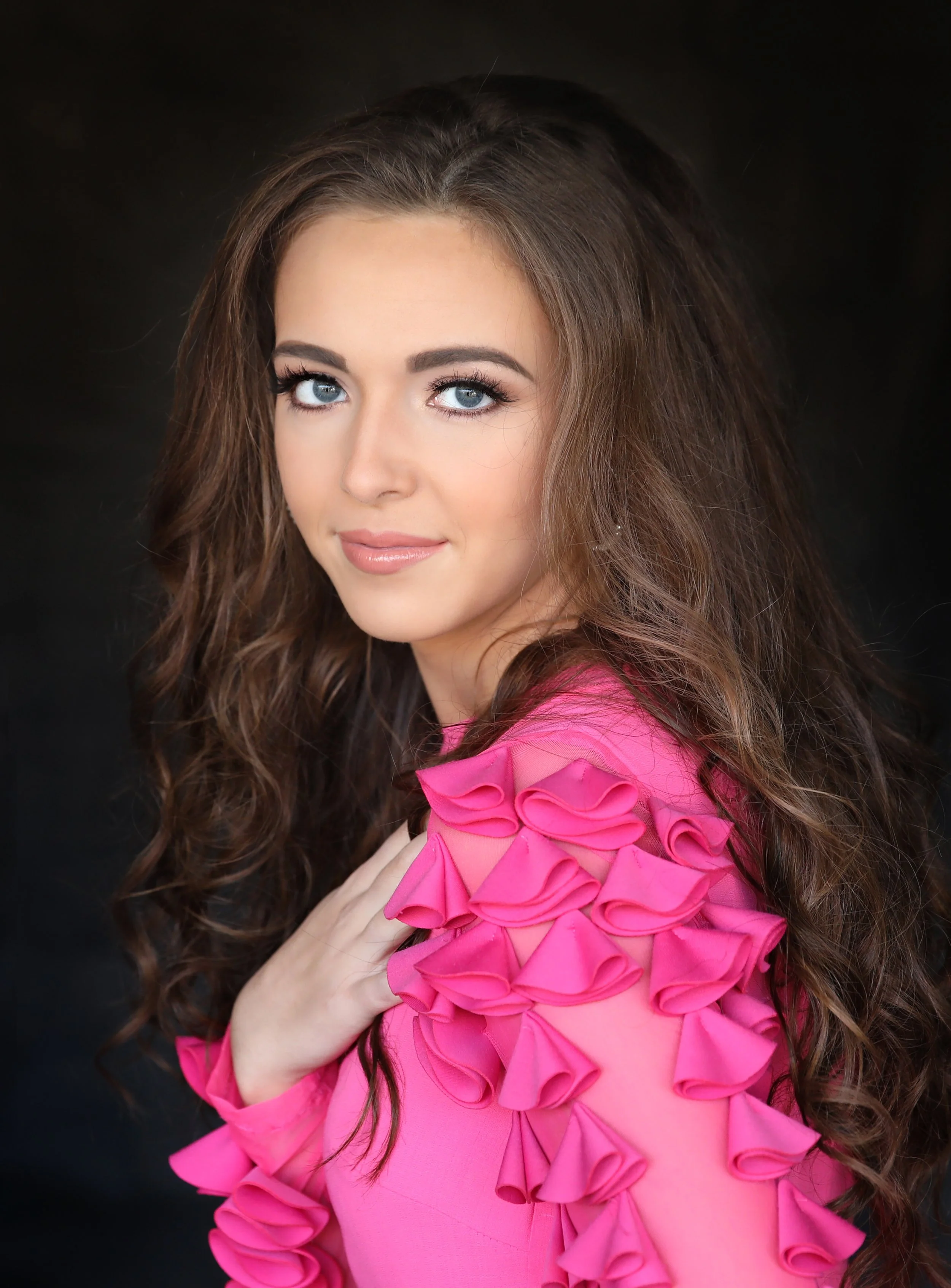 A young woman with long, curly brown hair, blue eyes, and light skin, wearing a bright pink dress with ruffled details on the shoulder, posing against a dark background.