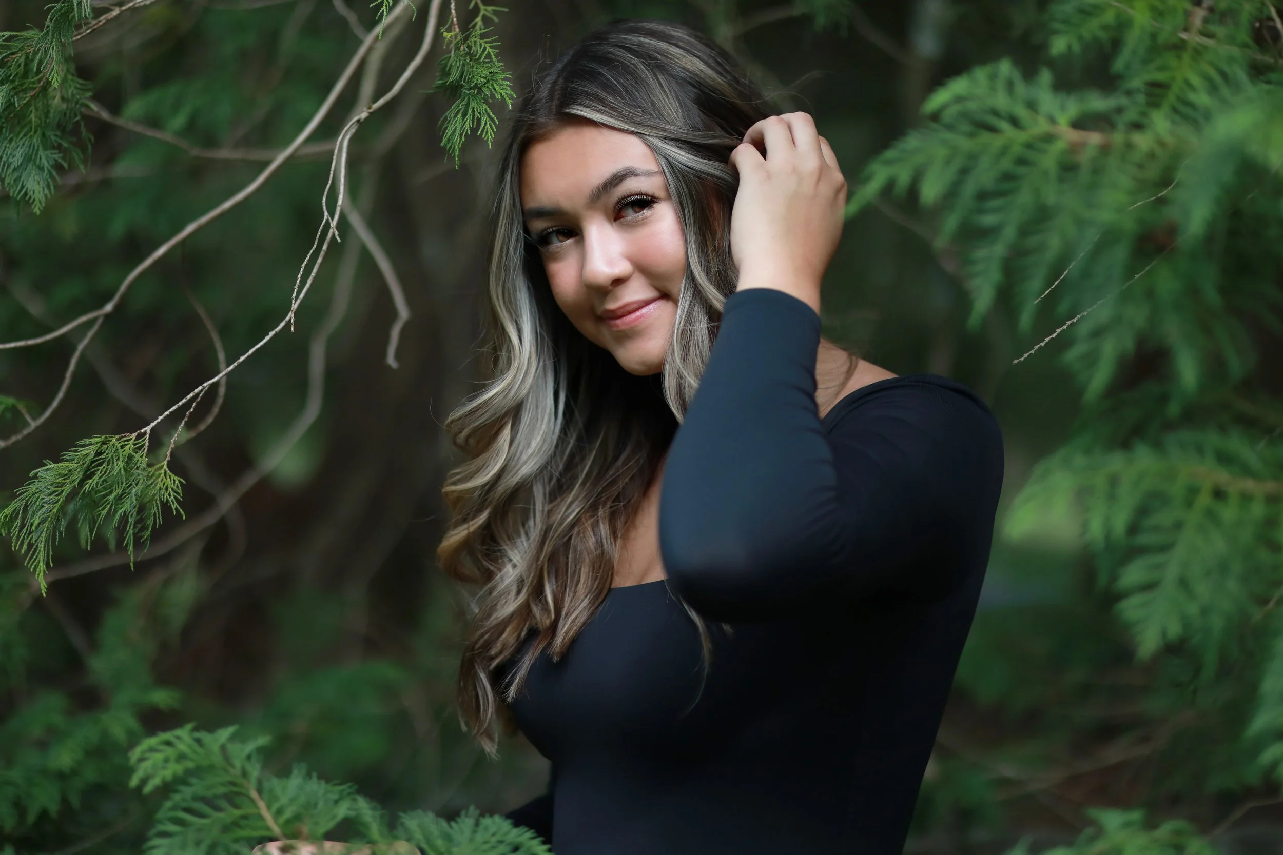 A woman with wavy, shoulder-length hair, wearing a black long-sleeve top, standing among green foliage, smiling and gently touching her hair.
