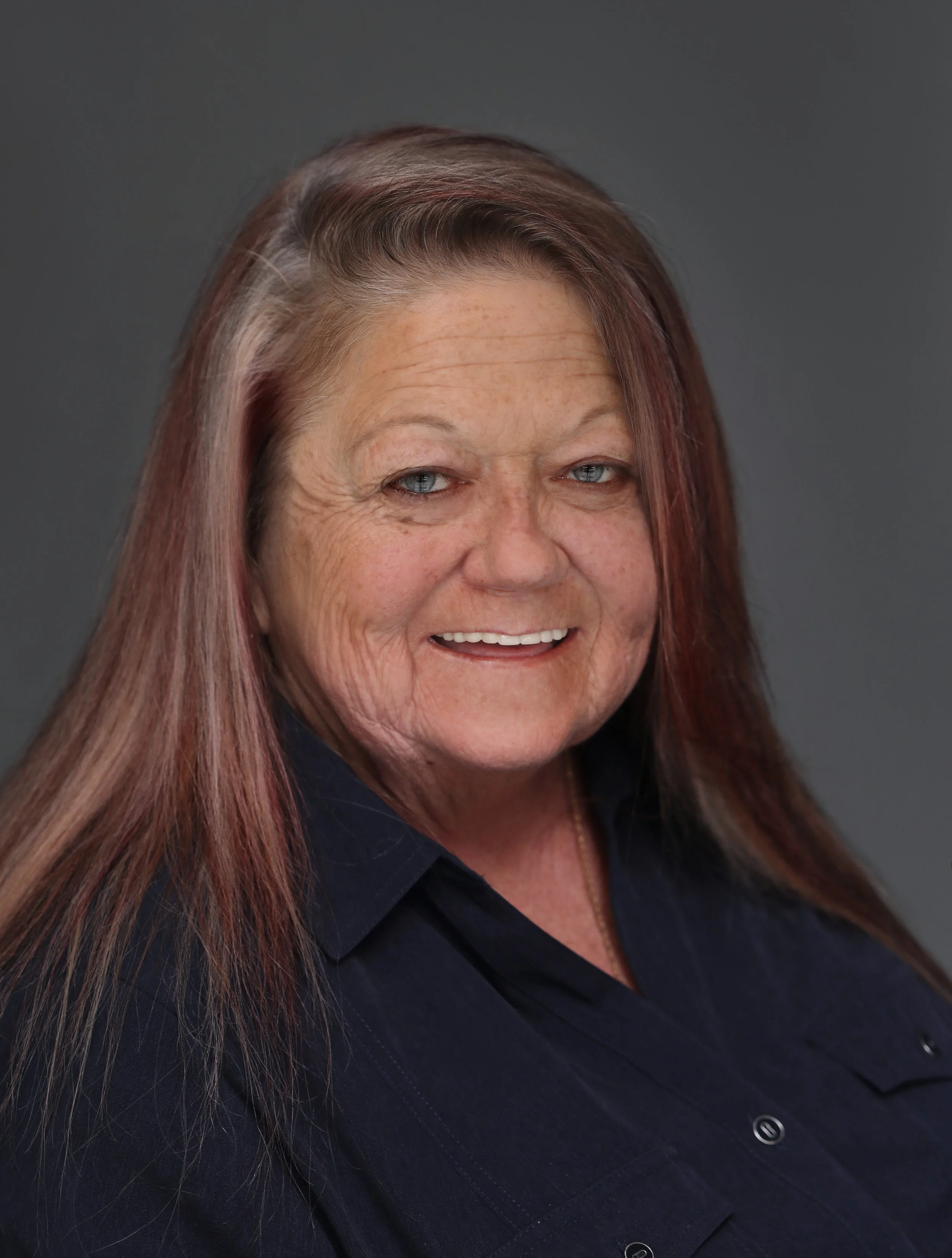 A smiling woman with long, reddish-brown hair, wearing a dark collared shirt against a plain gray background.