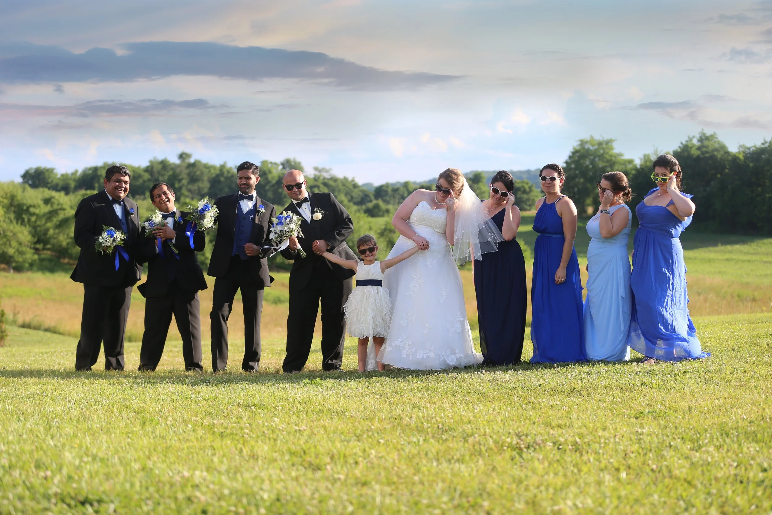 Wedding party standing outside on a grassy field, with some guests wearing sunglasses and smiling.