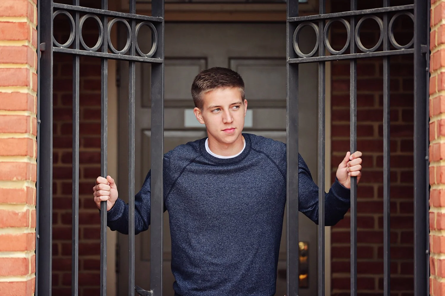 A young man standing behind a barred gate at a house entrance, holding the bars with both hands, looking to the side with a thoughtful expression.