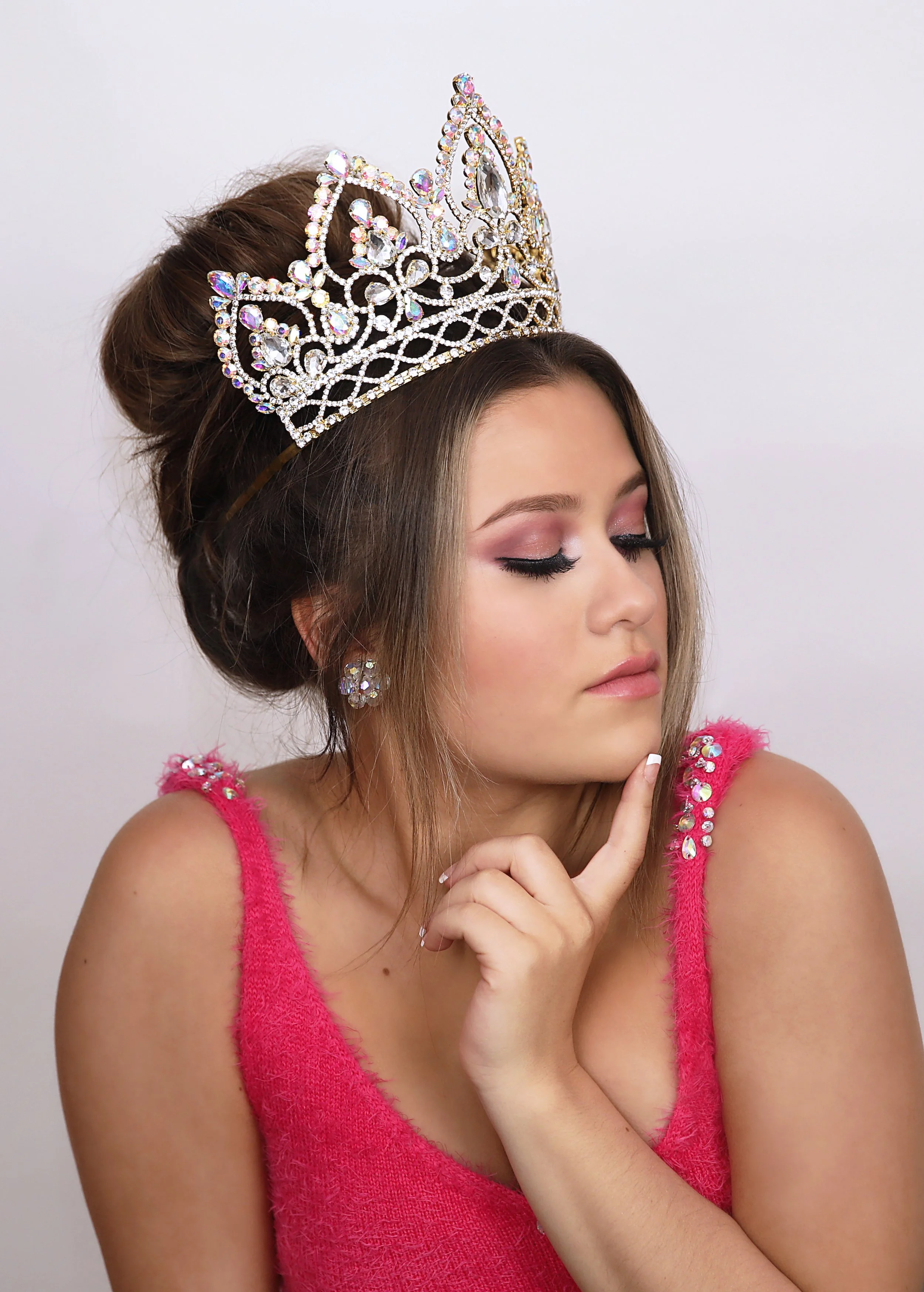 Young woman wearing a crown and pink dress, with makeup and jewelry, posing with her eyes closed and finger on her chin.