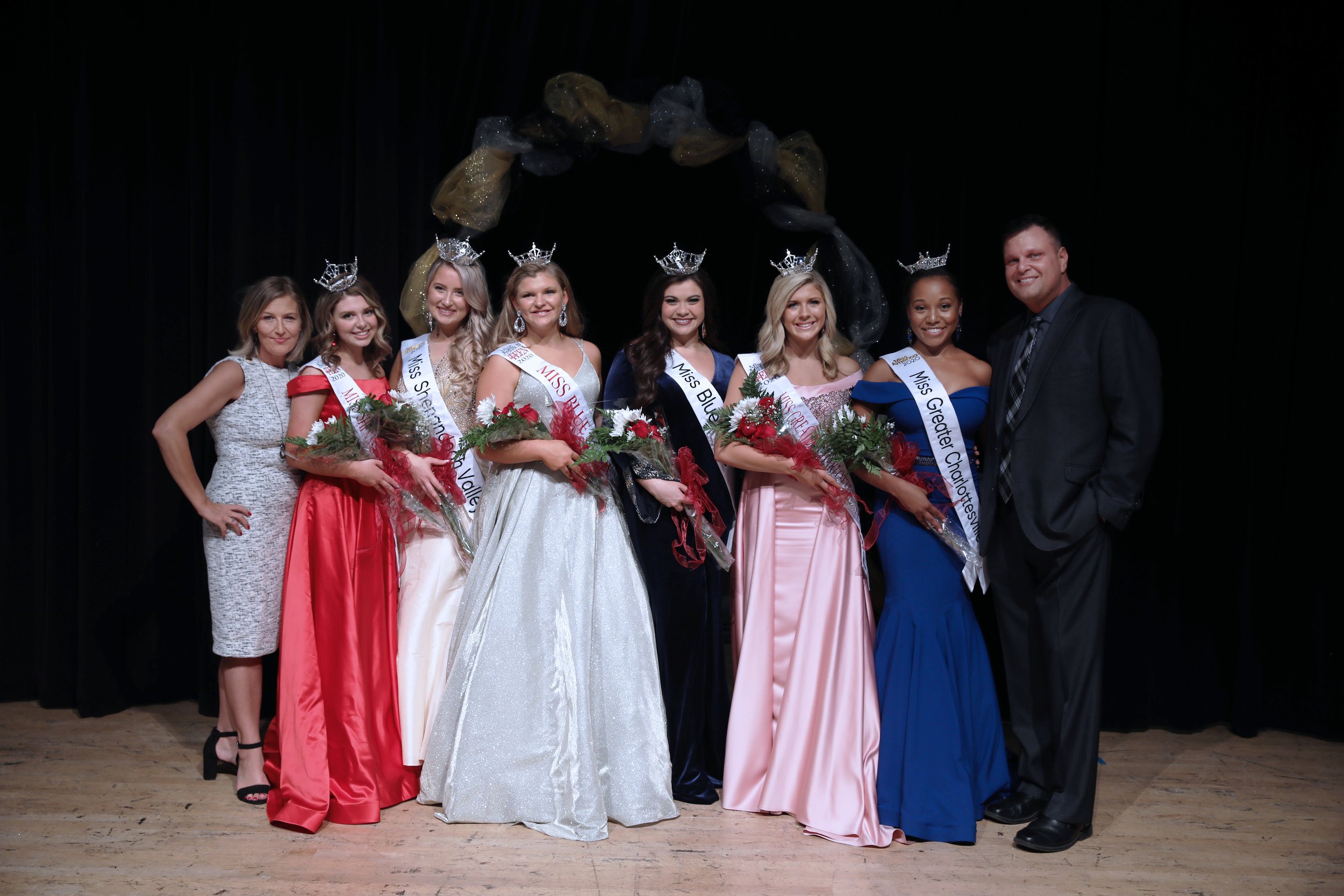 A group of seven women in formal dresses and crowns, holding bouquets of flowers, stands on a stage with a black curtain backdrop, celebrating a pageant or crowning event, accompanied by a man in a suit.