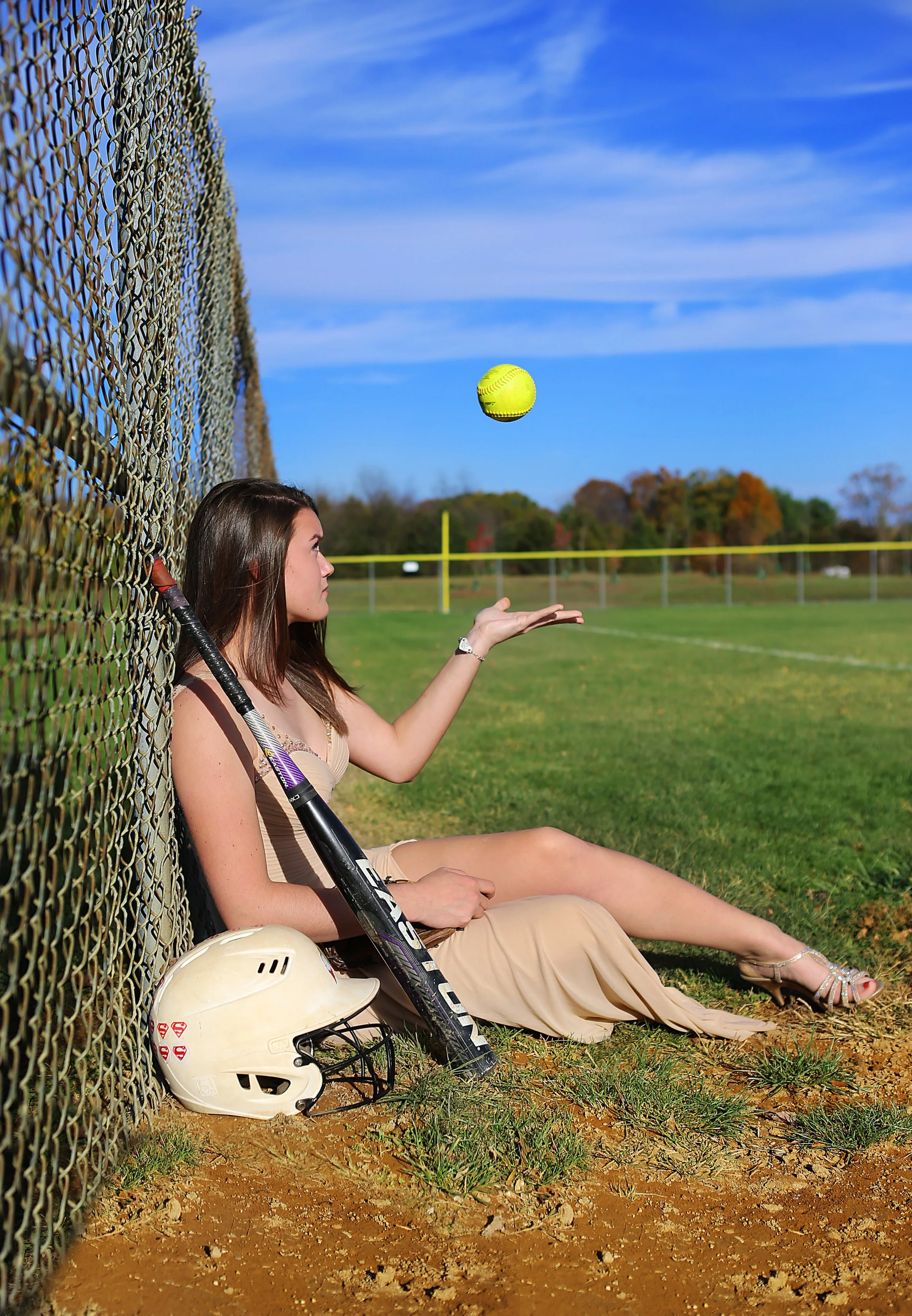 A woman sitting on the ground next to a baseball fence, holding a baseball bat, with a notice helmet and a tennis ball in the air.