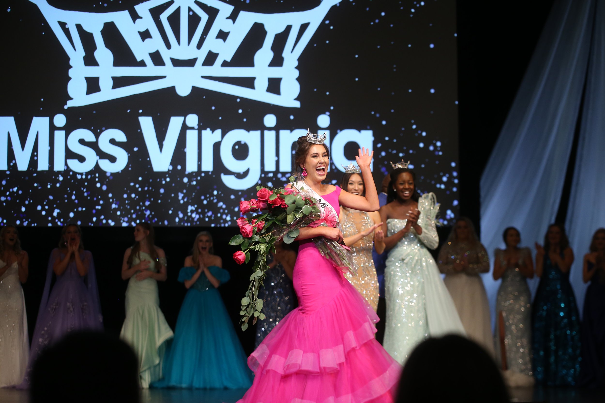 Miss Virginia crowned in a pink gown holding a large bouquet of pink roses on stage, with other contestants in elegant gowns clapping and celebrating behind her, and a large screen displaying 'Miss Virginia' in the background.
