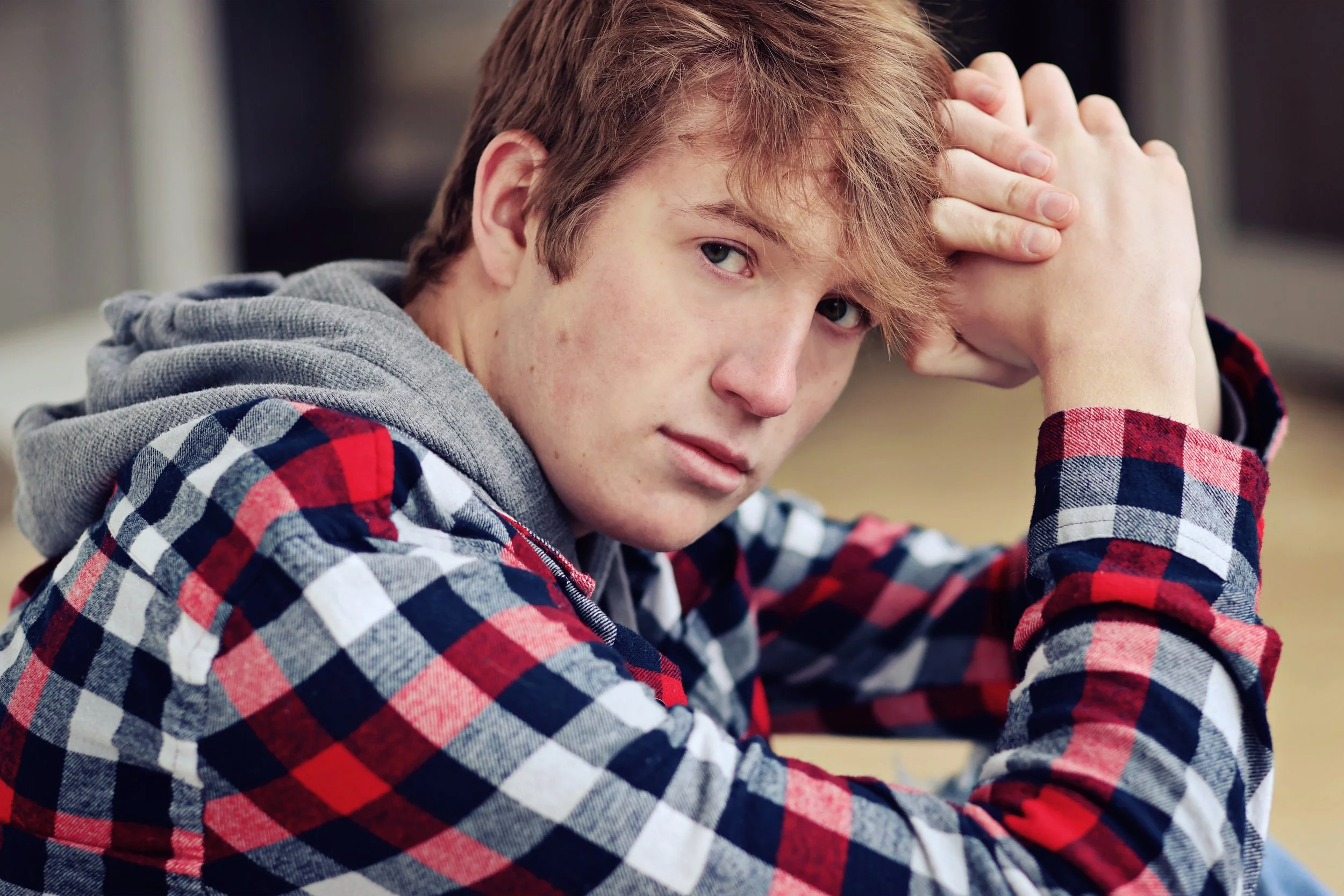 A young man with reddish hair, wearing a red and black checkered shirt and a gray hoodie, resting his head on his hand in a contemplative pose.